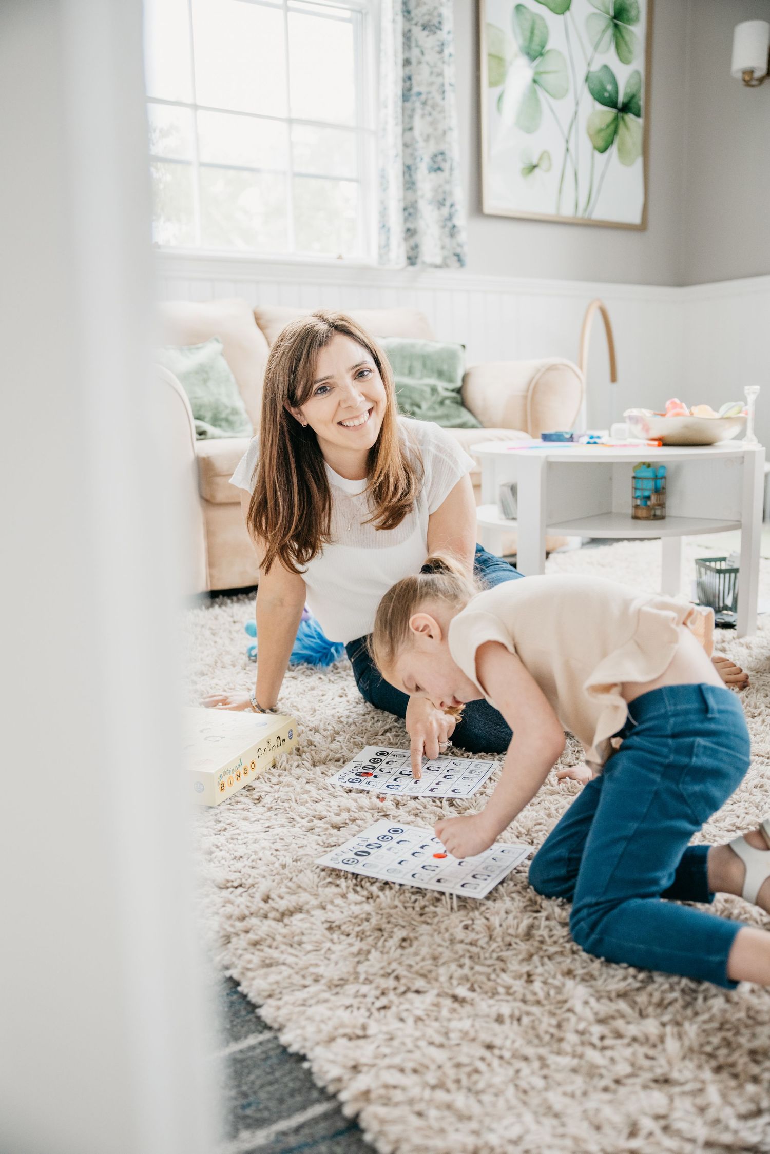 A smiling mother watches her young child work on a puzzle board on a cozy carpet in a bright, cheerful living room.
