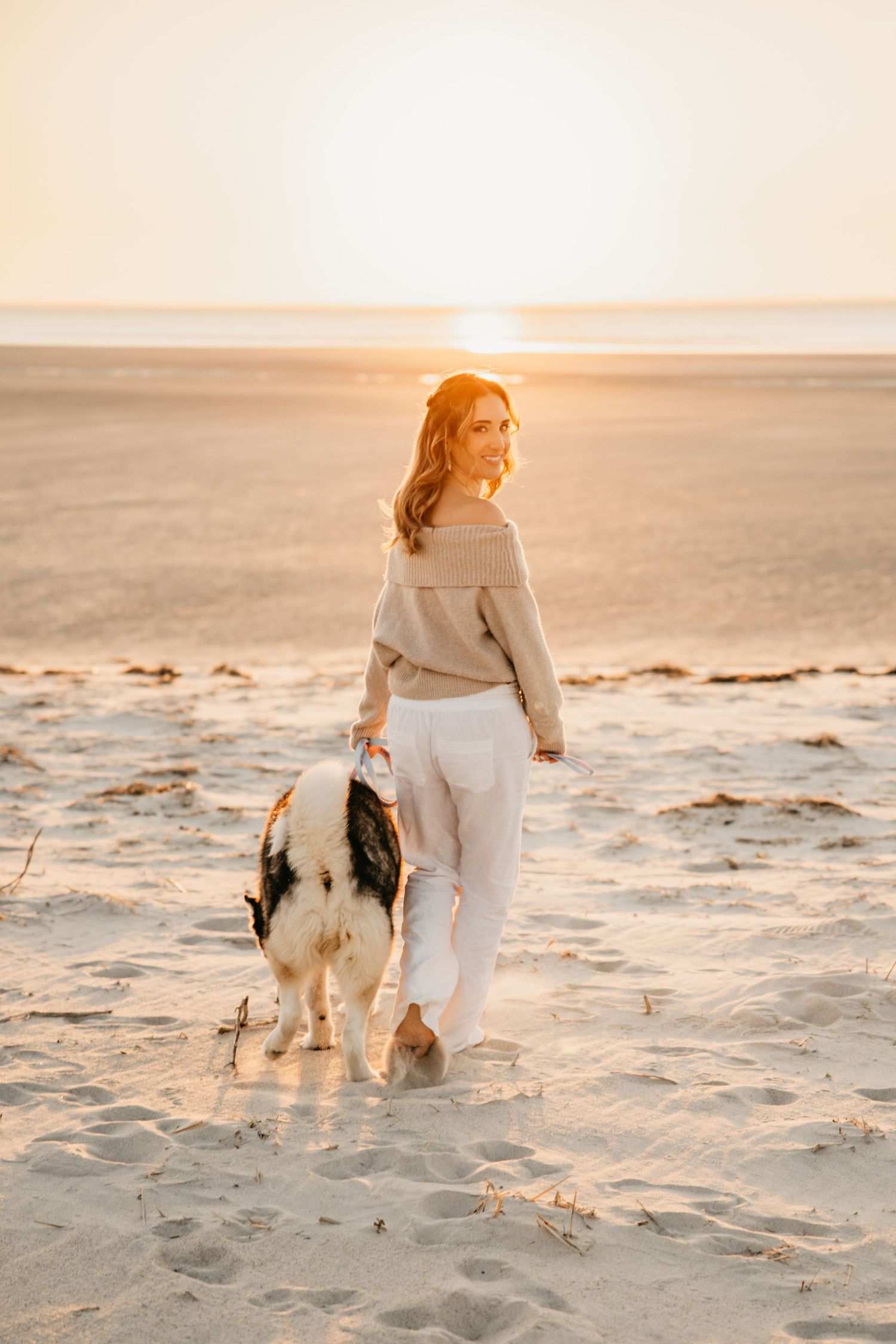 Woman walking dog on sandy beach at golden sunset, wearing beige sweater and white jeans.