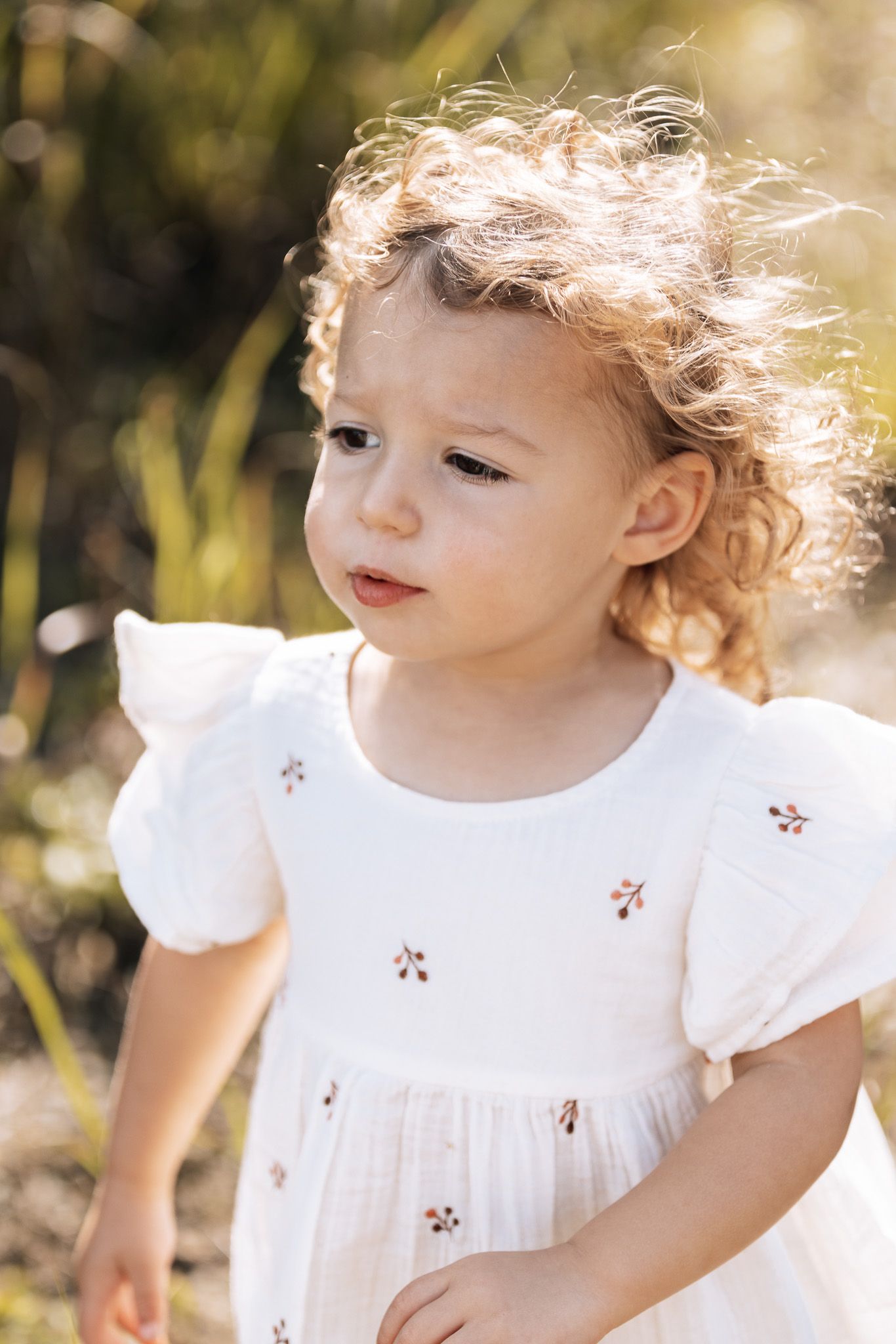 A child in white dress with curly hair illuminated by golden sunlight outdoors.