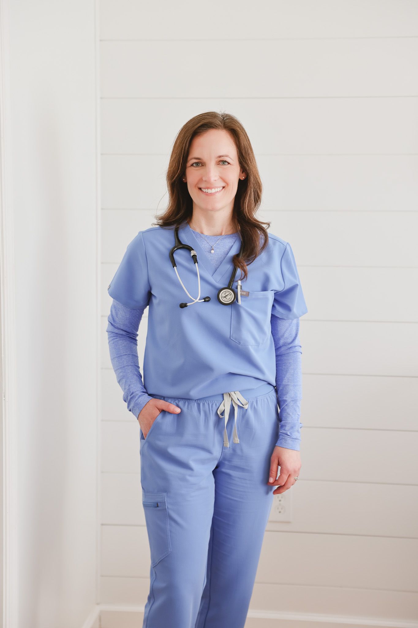 Smiling female healthcare professional in light blue scrubs with stethoscope standing against white shiplap wall.