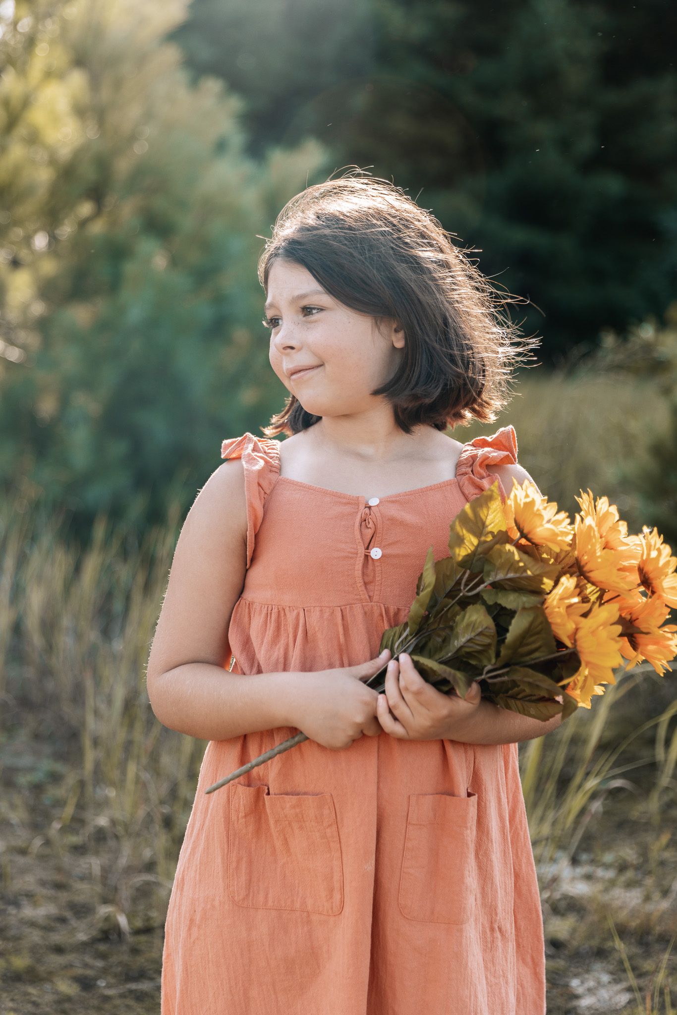 A child holds vibrant sunflowers while wearing a coral-colored dress outdoors.