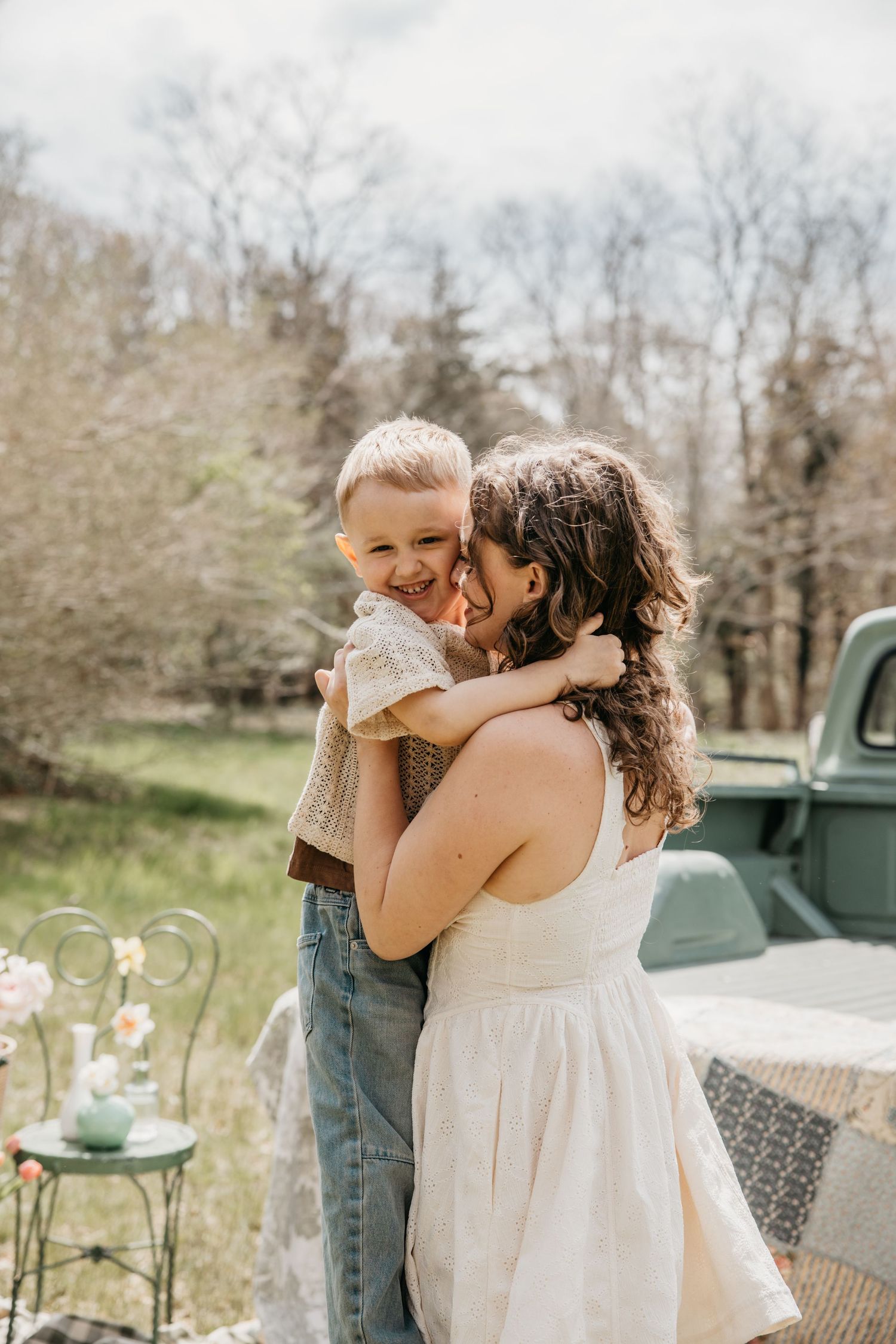 A woman in a white dress hugs and kisses a laughing young boy outdoors near a vintage green truck in spring.