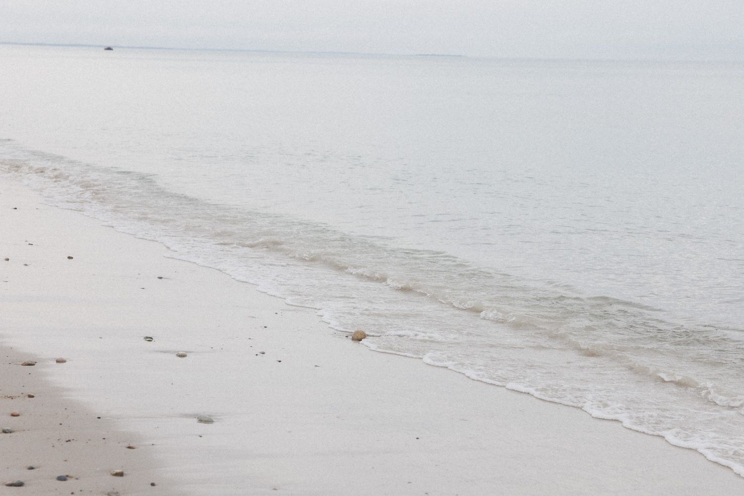 Gentle waves wash up on a misty white sand beach under an overcast sky.