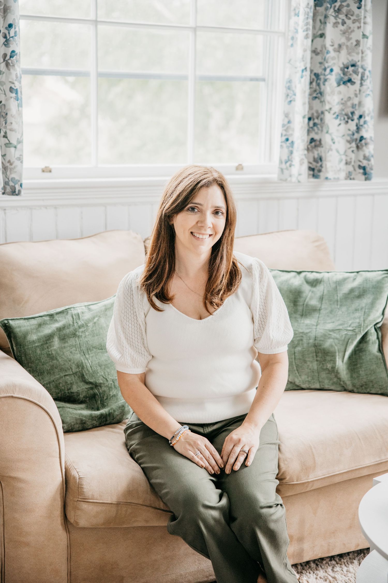 Smiling woman with brown hair sits on beige sofa with green pillows, wearing white top and olive pants.