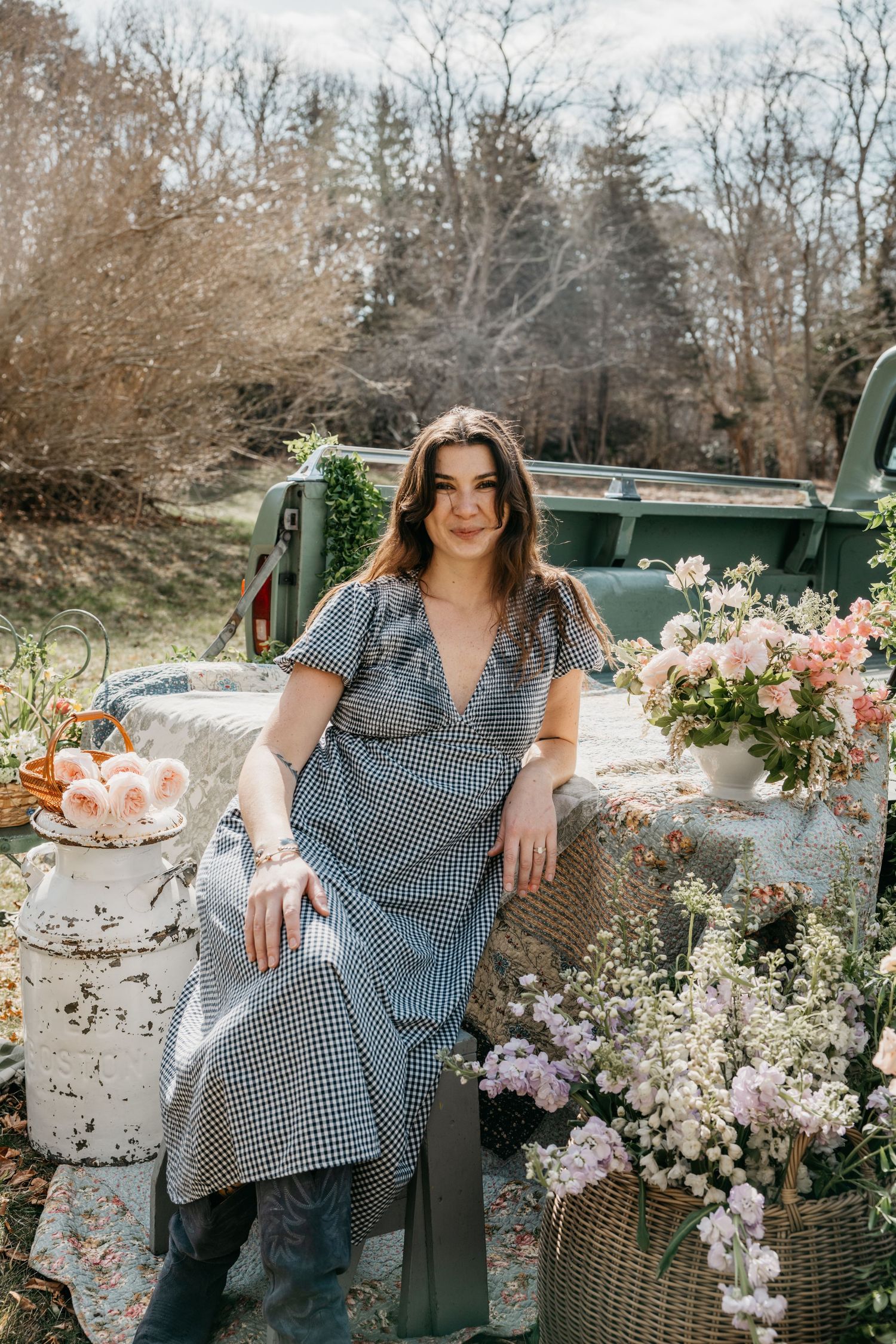 Woman in gingham dress poses among spring flowers by vintage green truck in outdoor setting.