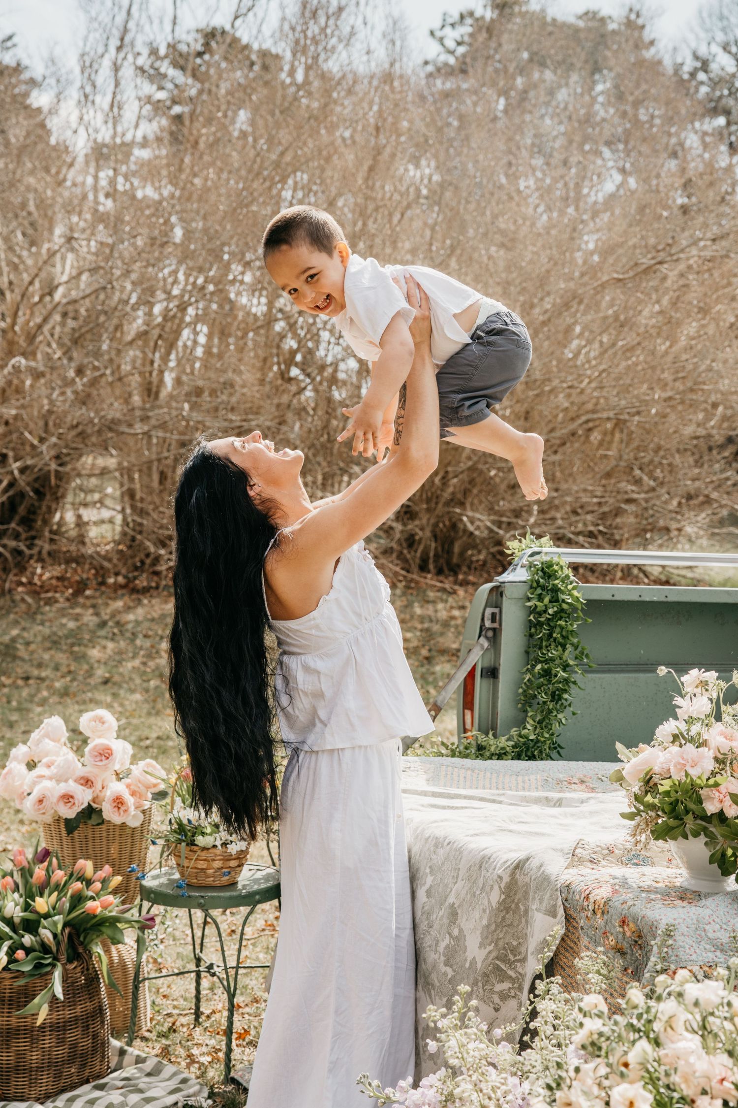 A joyful mother in a white dress lifts her smiling baby outdoors amid beautiful floral arrangements in soft pink tones.