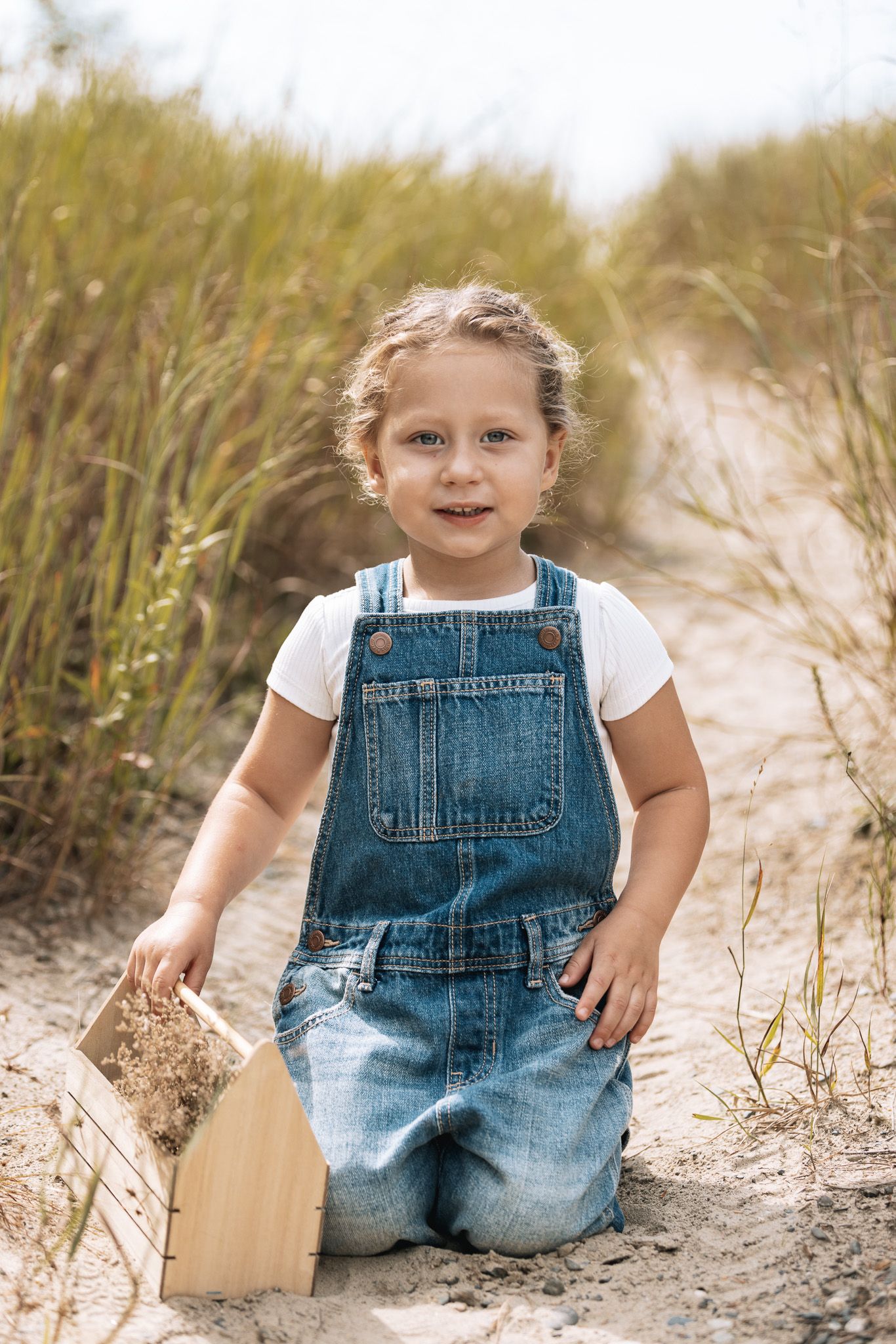 Child in denim overalls and white shirt plays on sandy ground near tall grass.
