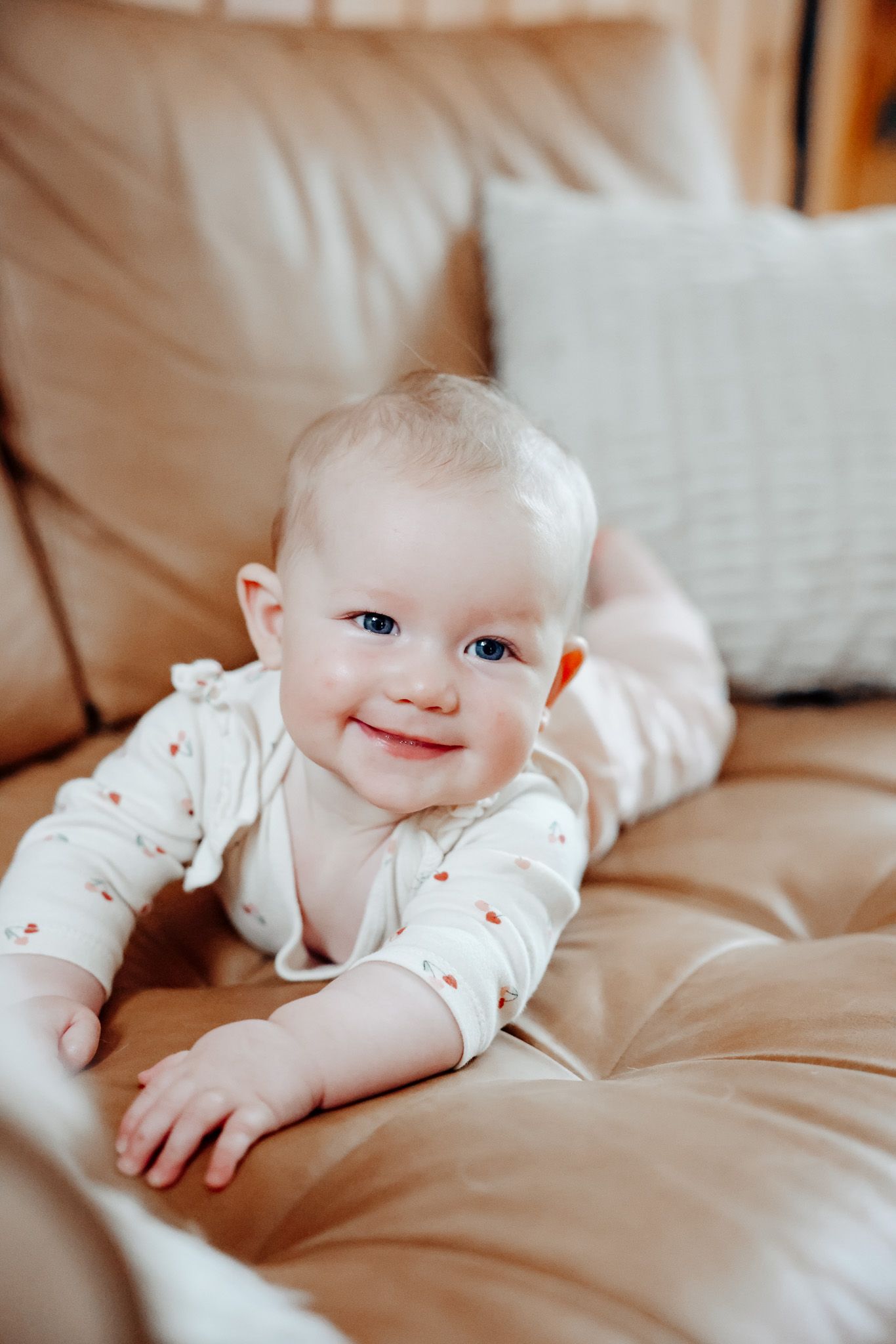 Baby in white floral outfit smiling while lying on beige couch.