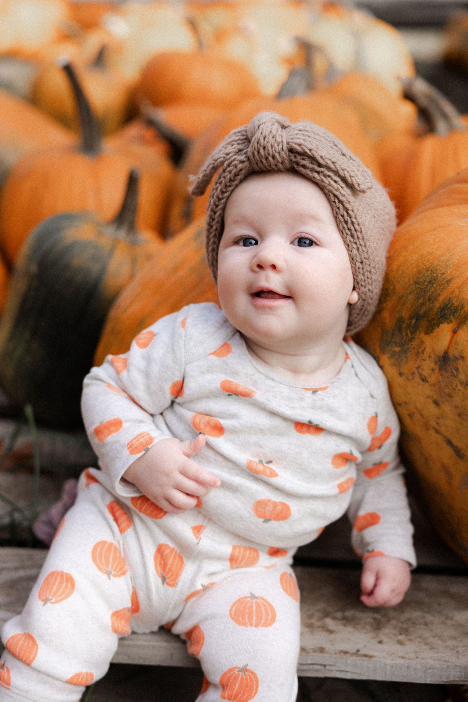 Baby wearing pumpkin-print pajamas sits among orange pumpkins.