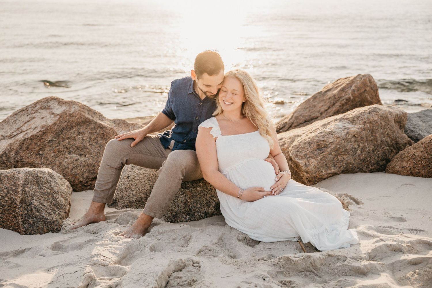Pregnant woman in white dress sits on beach with partner leaning on rocks during golden hour maternity photoshoot.