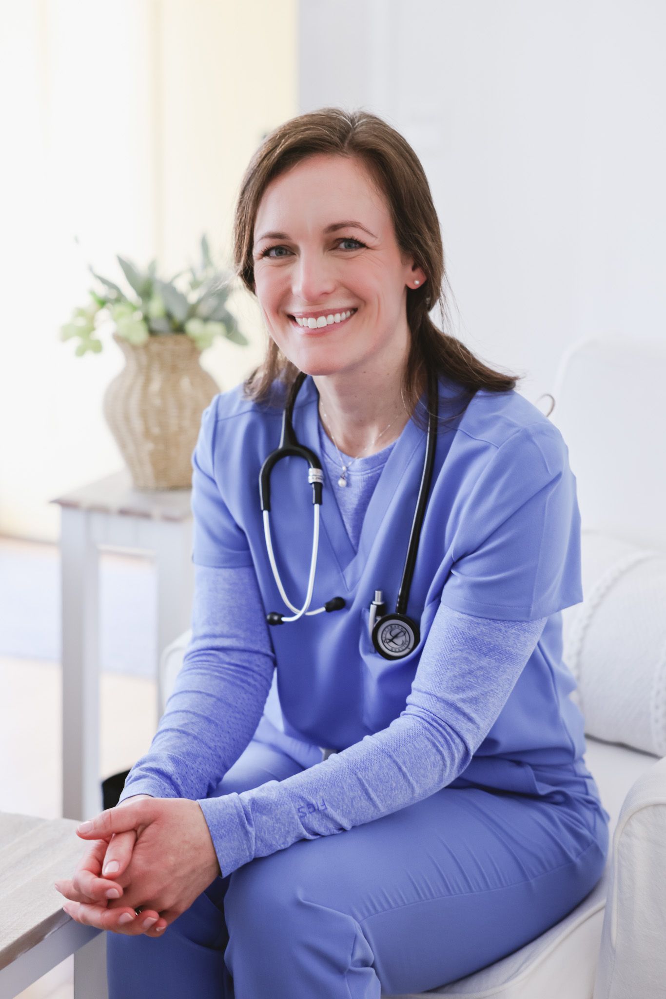 Smiling female nurse in blue scrubs with stethoscope sitting in a bright medical office setting.