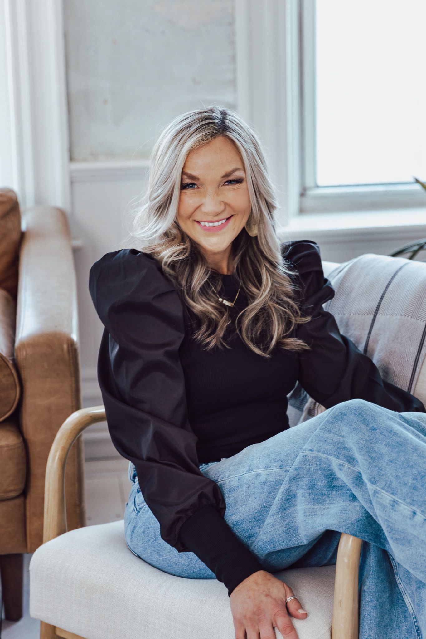 A woman in a black puff sleeve top and jeans relaxes on a grey couch in natural lighting.