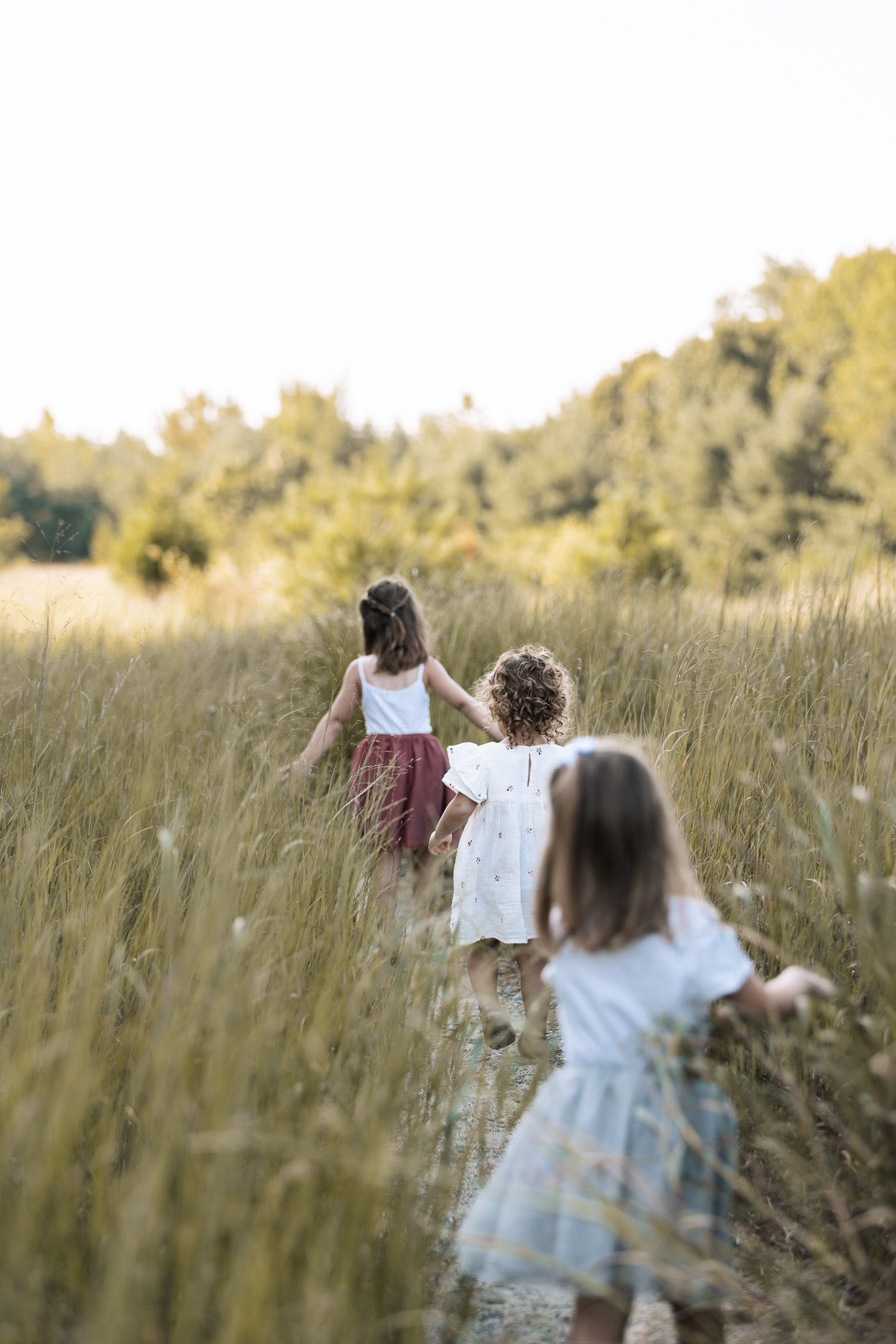 Children run through a sunlit meadow with tall grass and wildflowers.