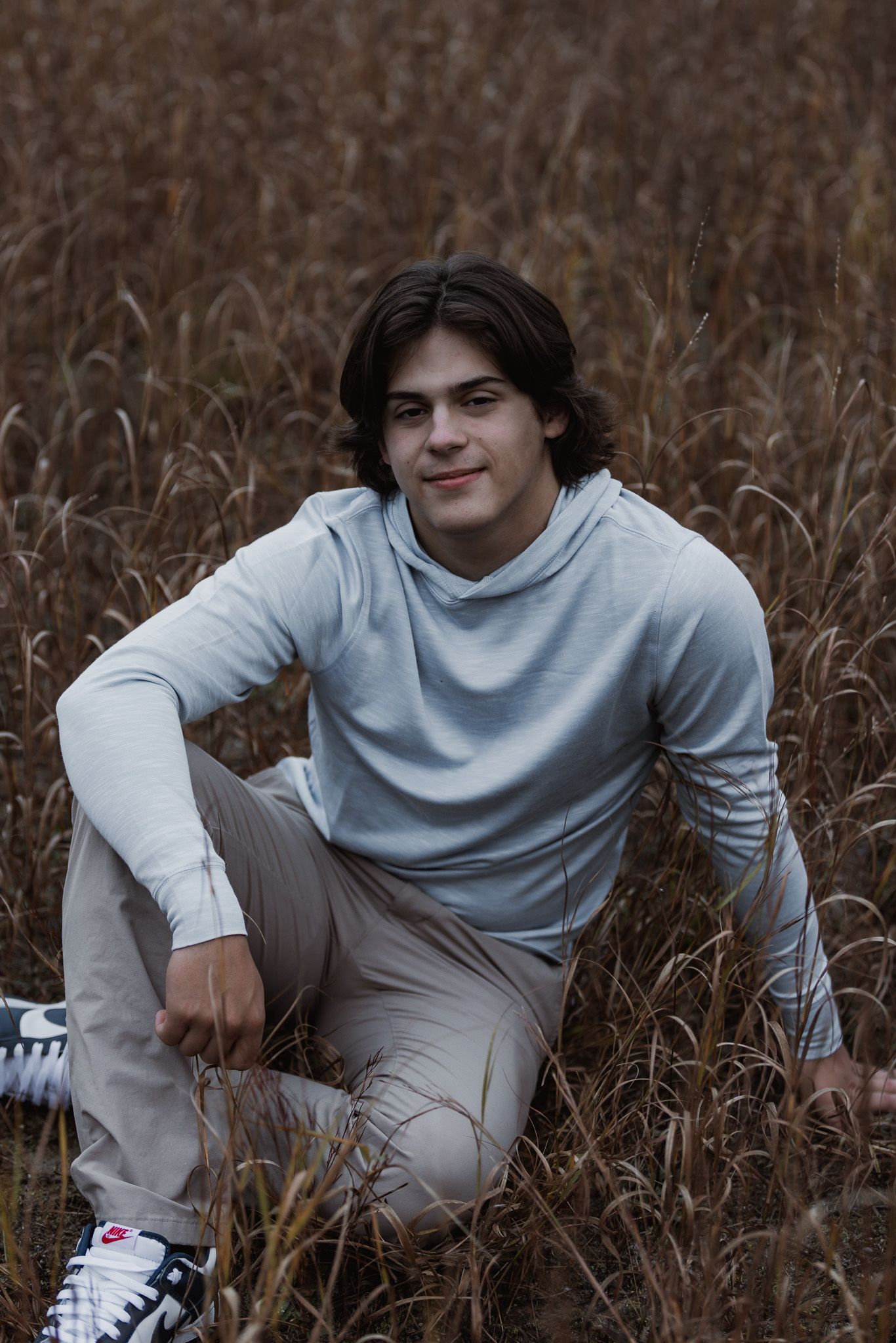 Person in light gray sweater poses casually in tall brown grass during outdoor photoshoot.