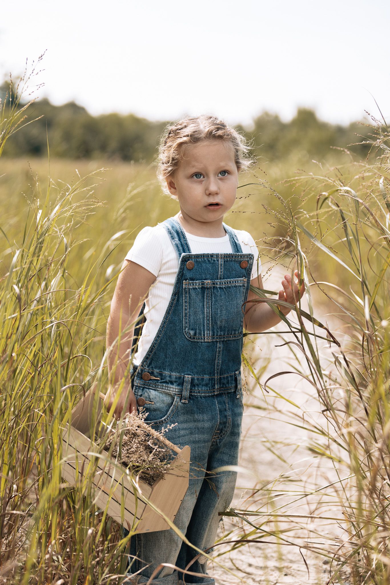 Young child in denim overalls explores tall grass in a natural outdoor setting during golden hour.