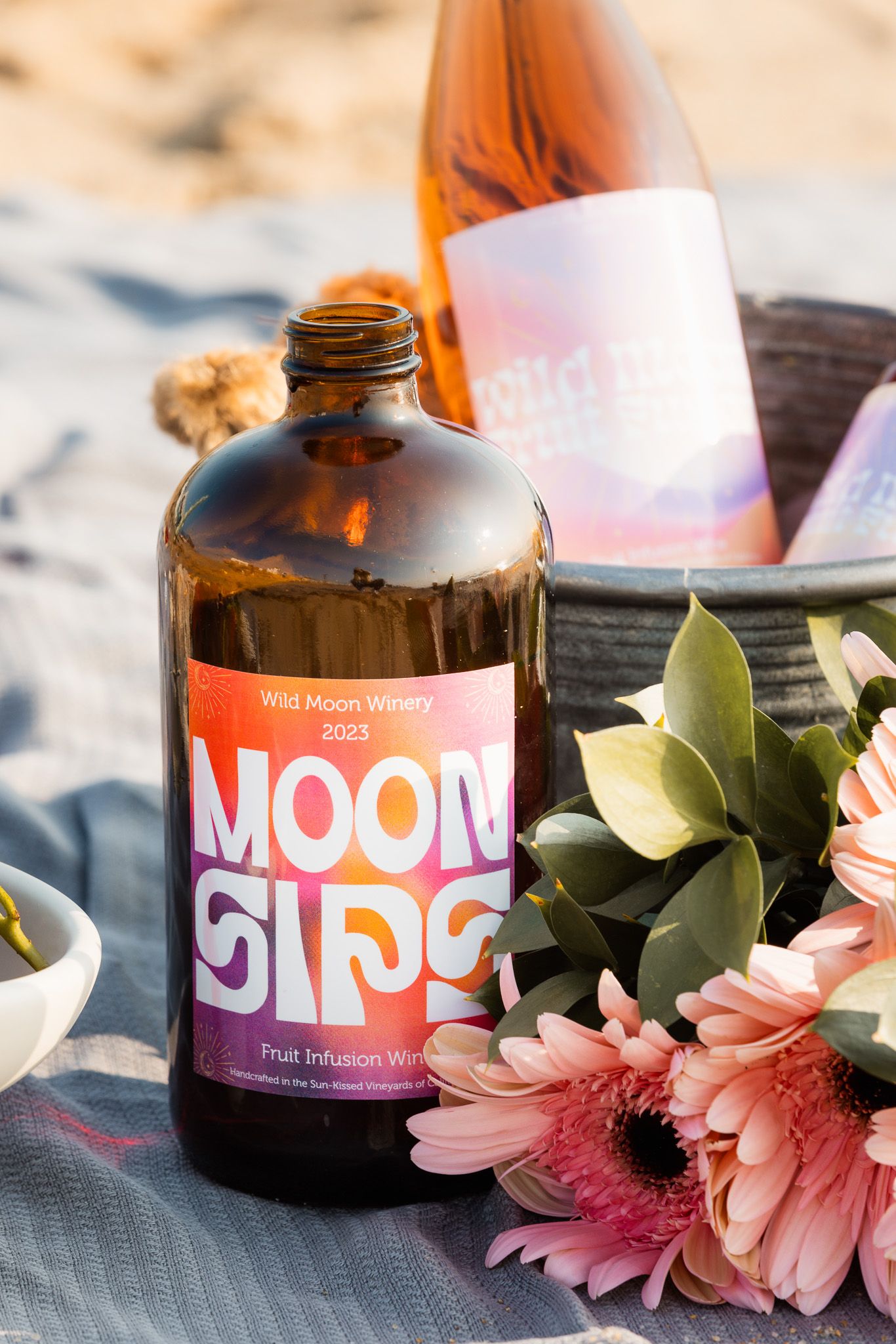 Moon Juice bottles arranged with pink flowers on beach blanket.