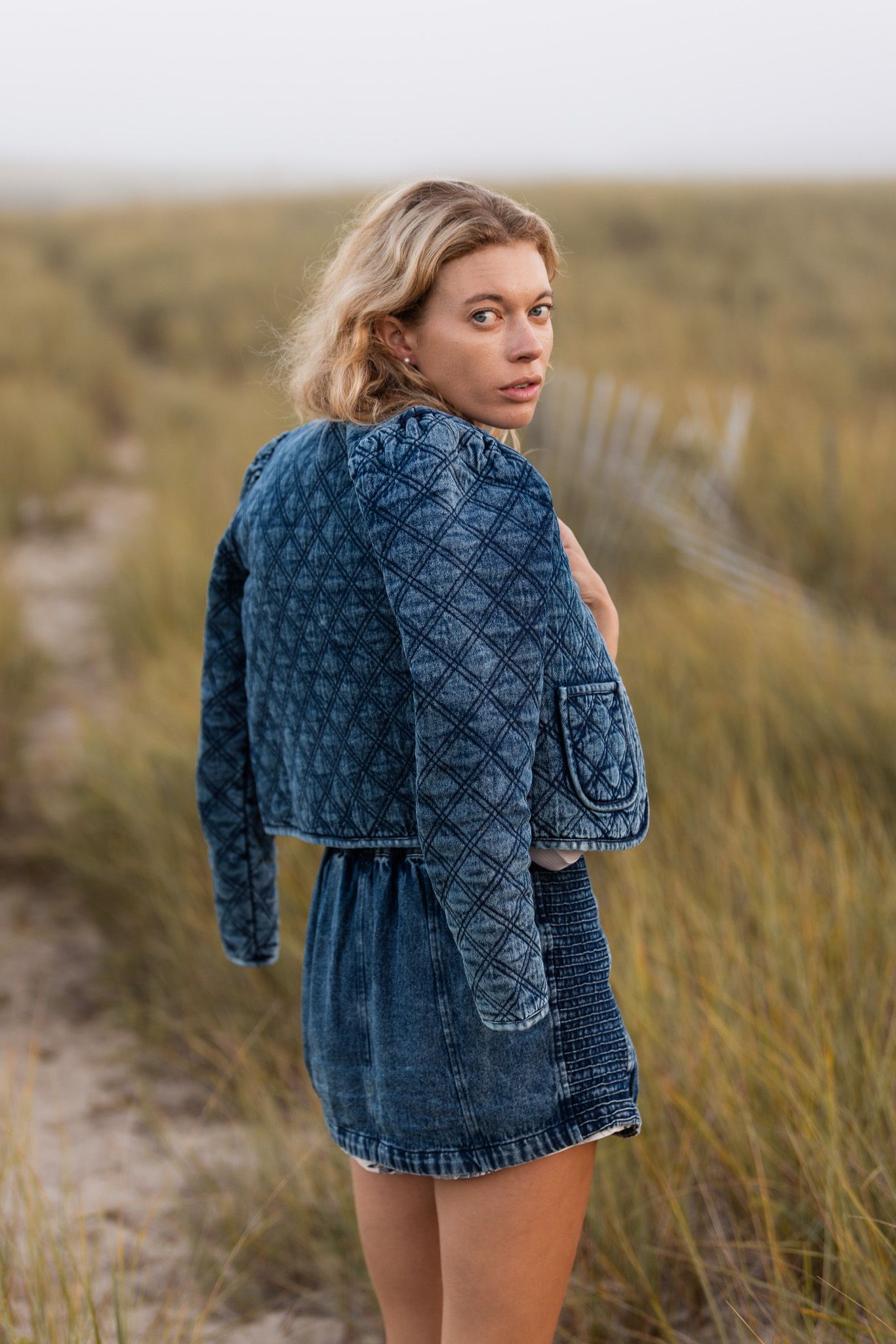 Woman in denim outfit looks over shoulder while standing in coastal grass.