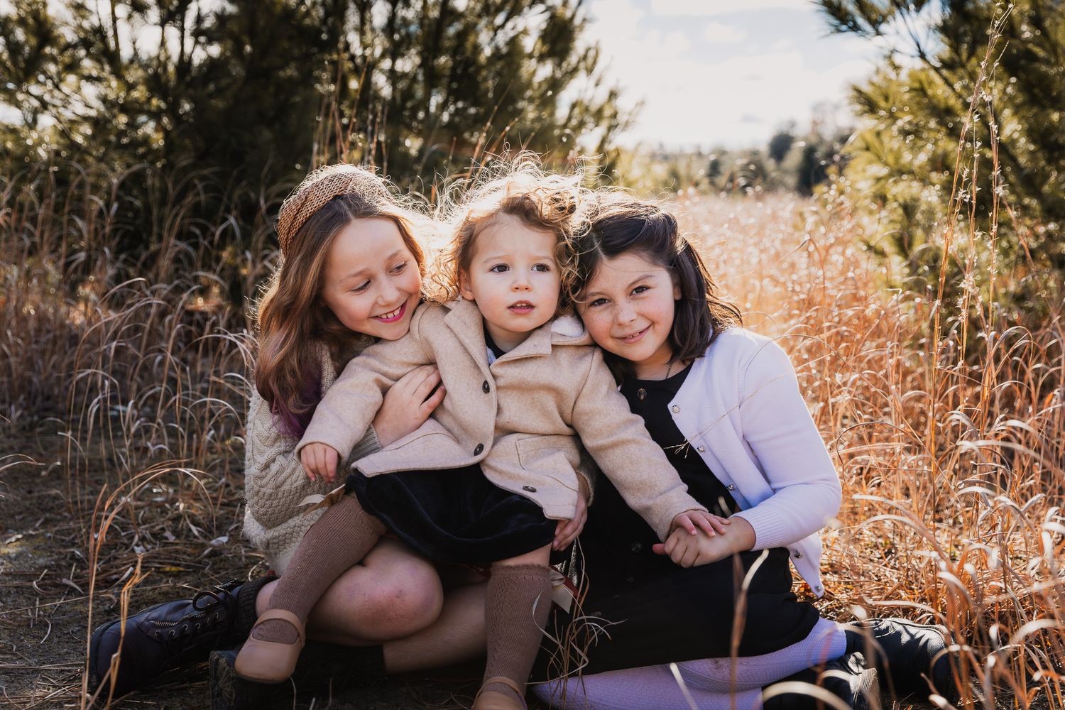 Three people share a tender moment outdoors among tall grasses at sunset.