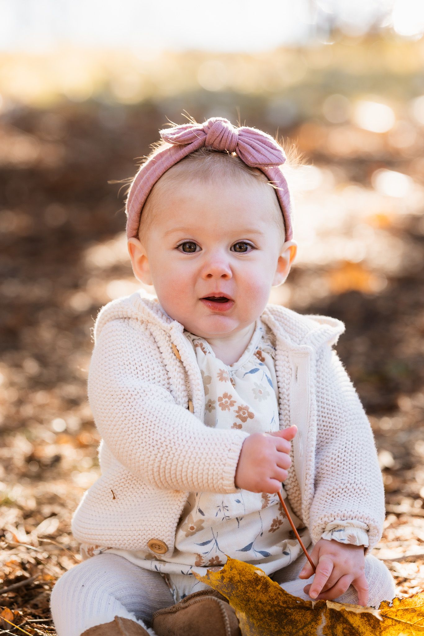Baby in white knit sweater sits on autumn leaves during golden hour.