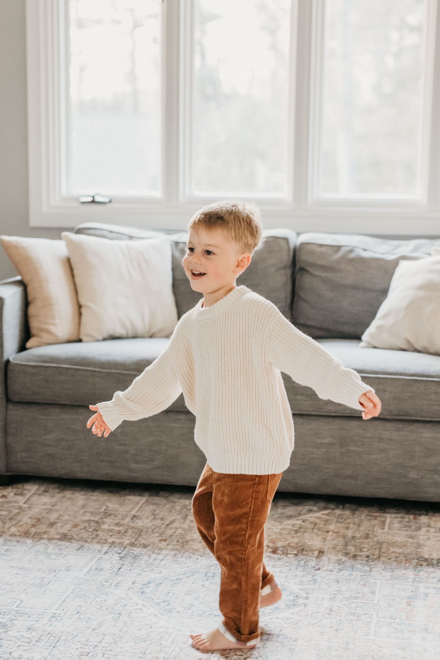 A small child in a cream sweater and brown pants dances playfully on a light colored carpet near a gray sofa.