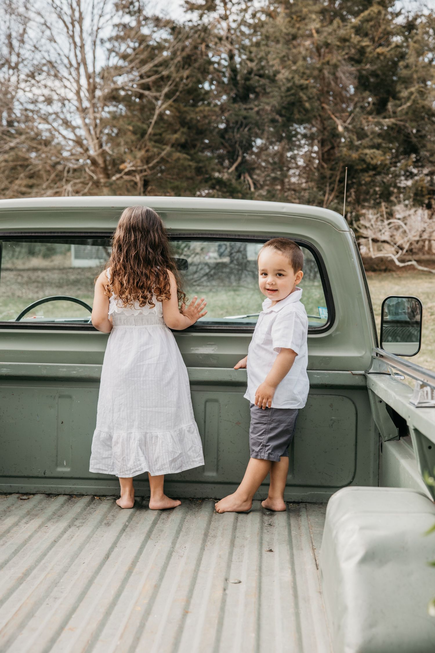 Two barefoot children in white stand on the bed of a vintage green pickup truck on a cloudy day.