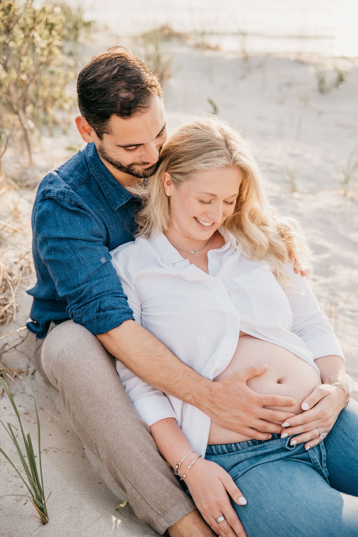 A couple sits on a sandy beach, the man embracing a smiling pregnant woman as they both cradle her baby bump.