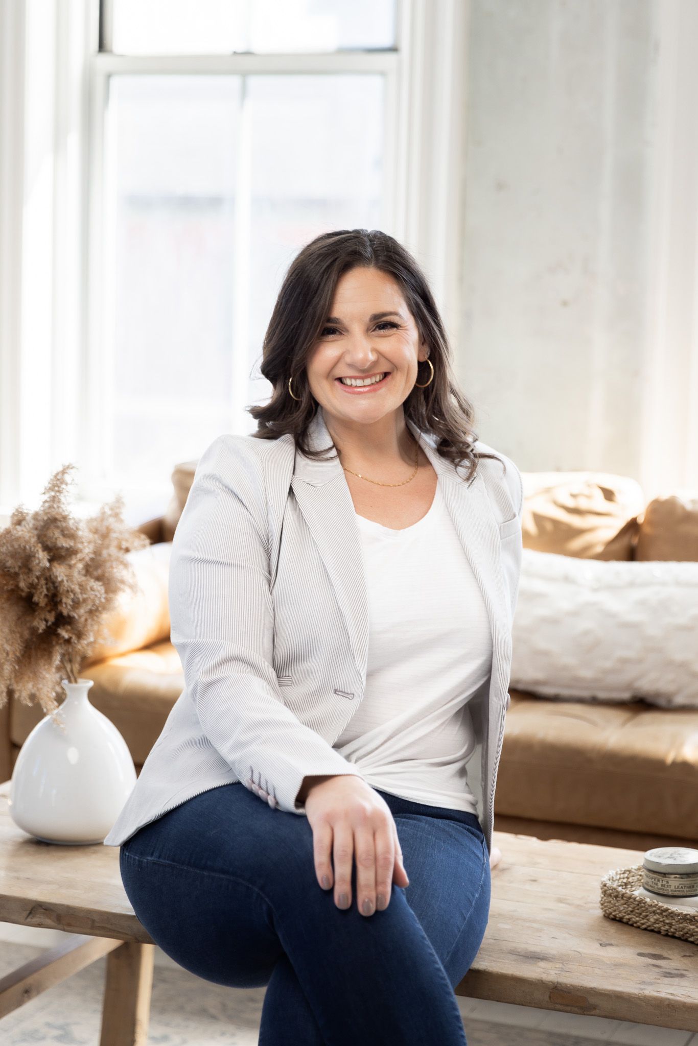 Person in white blazer and jeans sits casually in a bright, modern room with natural lighting.