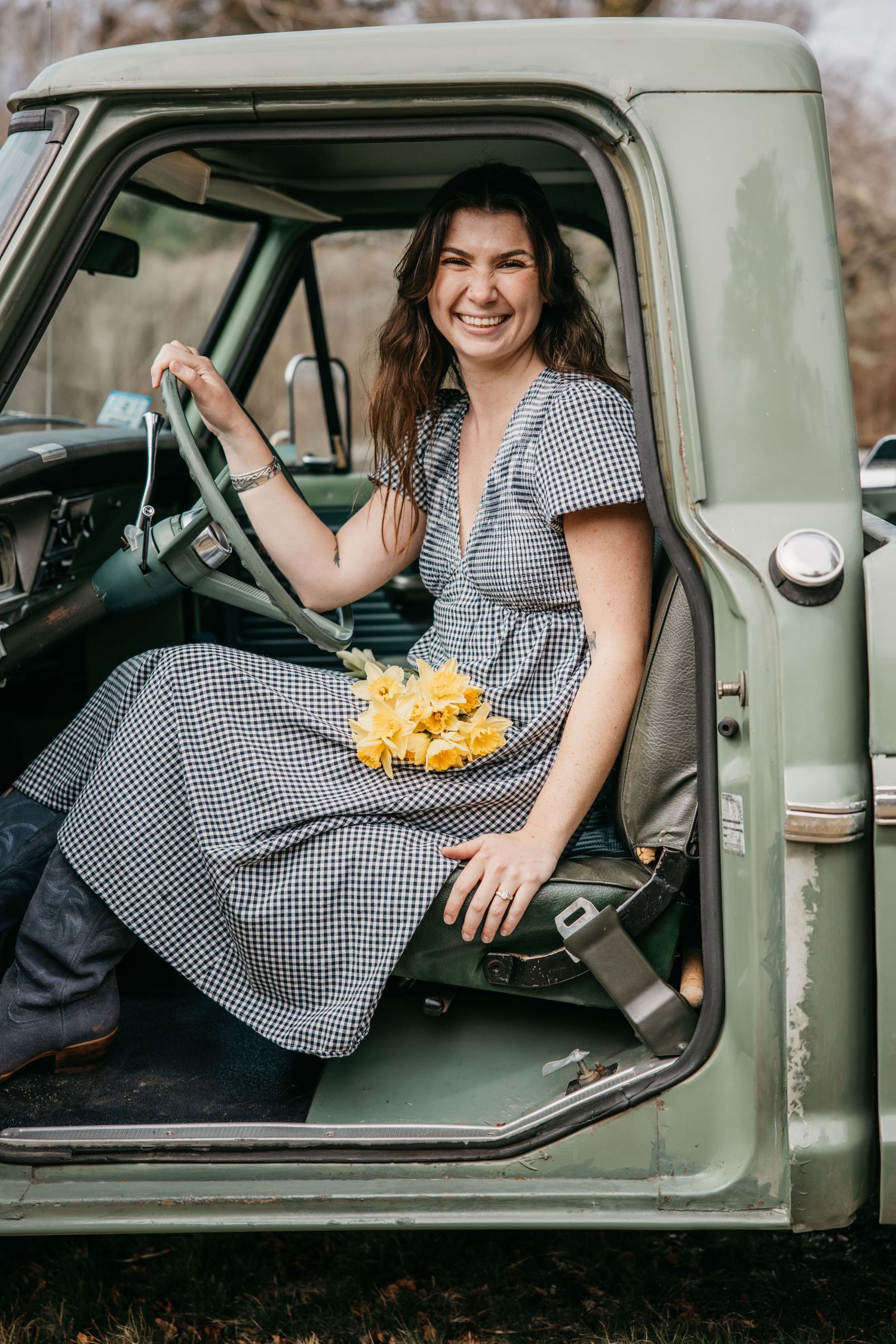 Smiling woman in gingham dress holding yellow flowers, sitting in vintage green truck with open door.