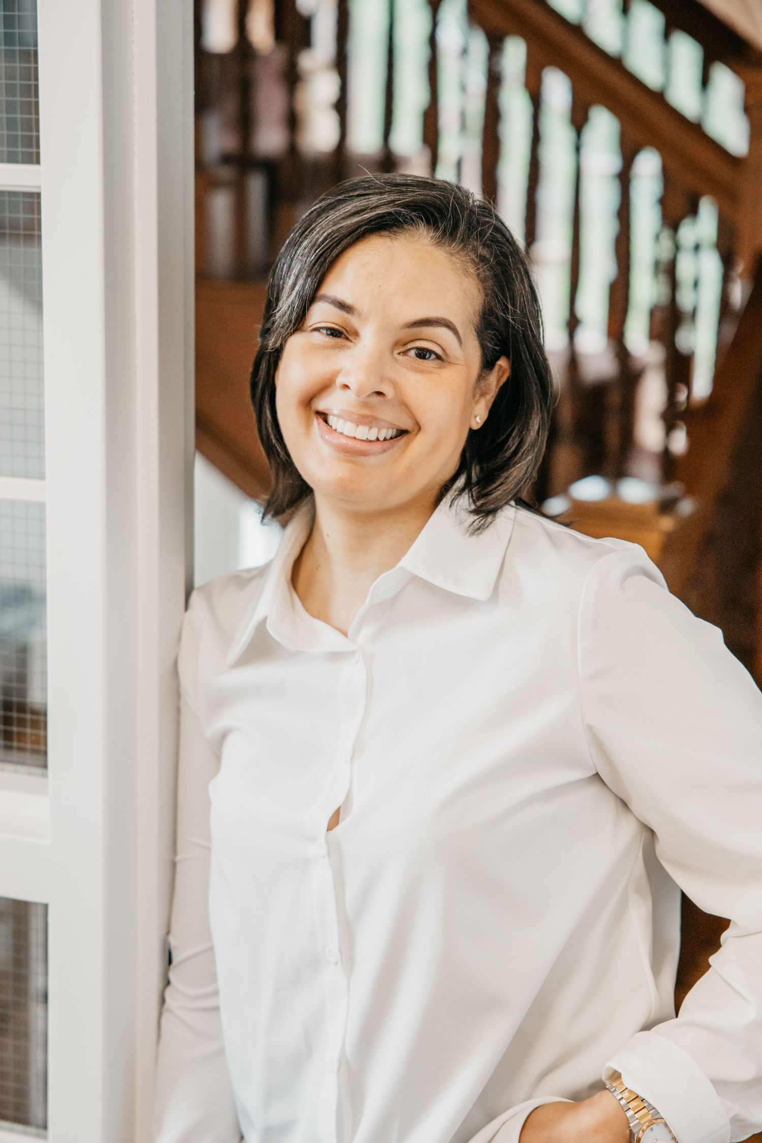 Smiling woman in white button-up shirt leaning against wall near staircase in bright indoor setting.