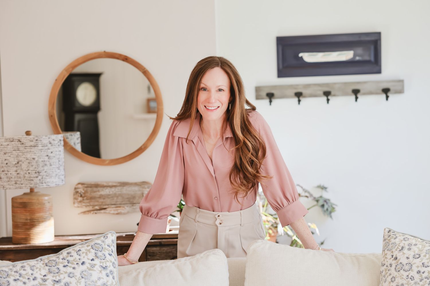 Smiling woman with long auburn hair wearing a pink blouse and beige pants leaning on a white sofa in a bright home.