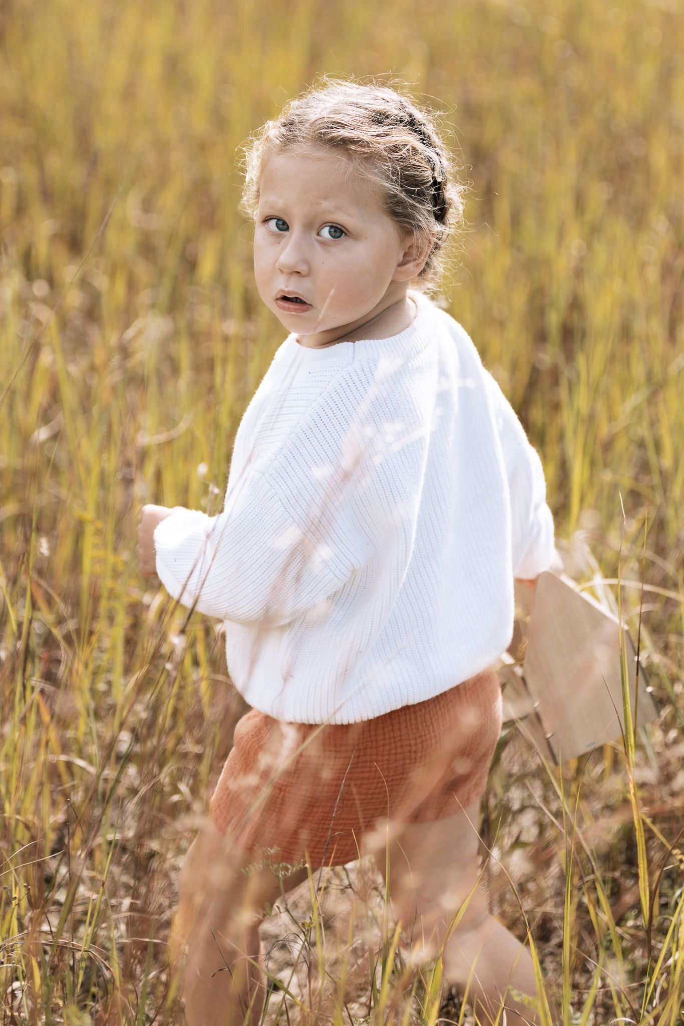 Child in white dress twirling through tall grass at golden hour.