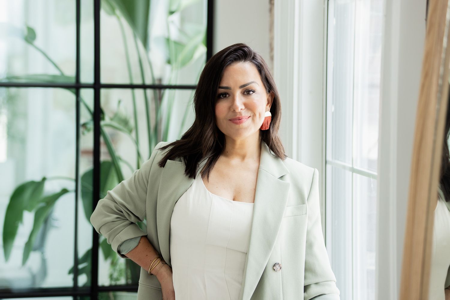 Person in light-colored blazer poses by large windows in a bright, modern space.