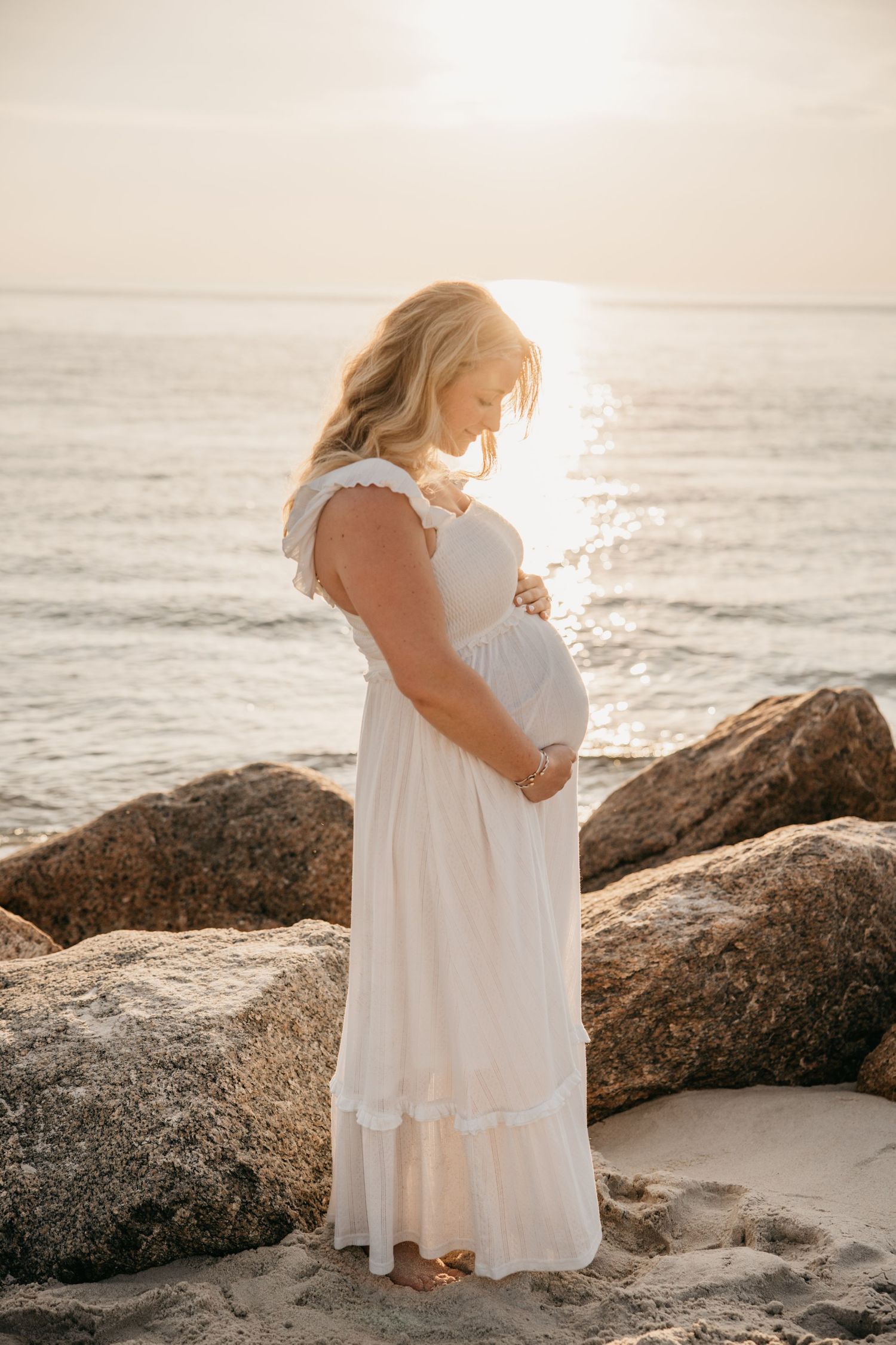 Pregnant woman in white dress cradling baby bump on rocky beach at golden sunset.