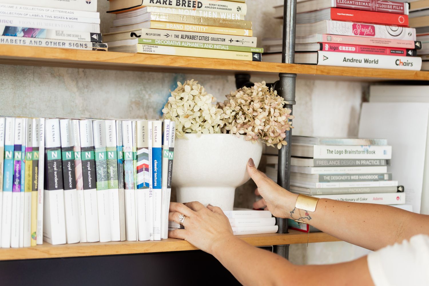 Hand reaches for books on wooden shelves filled with various titles and dried flowers.