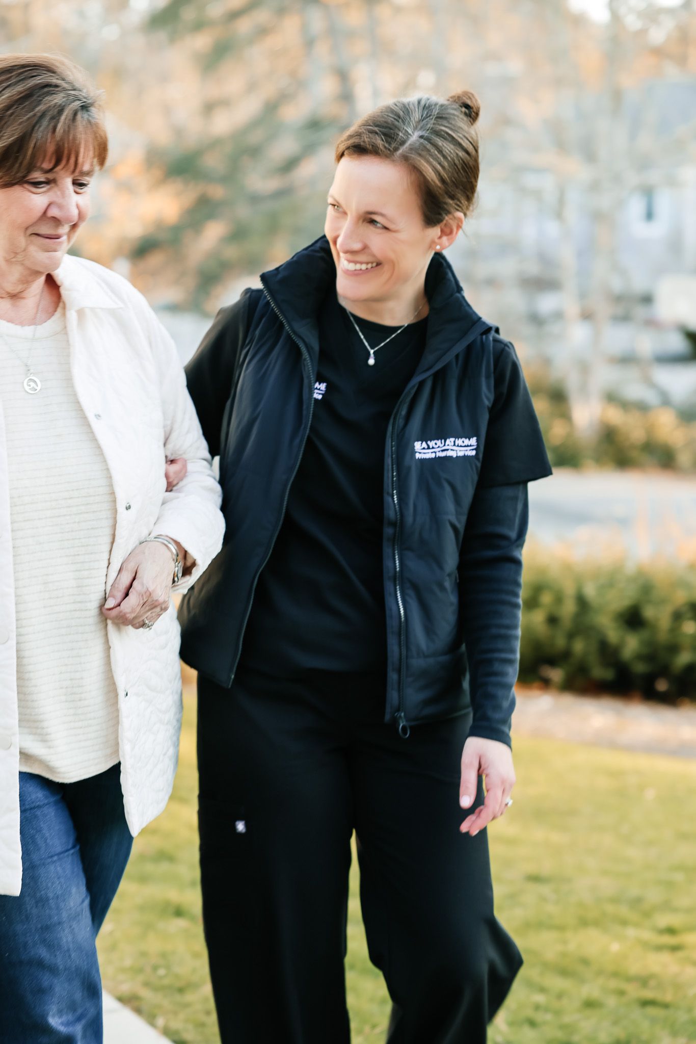 A caregiver in black uniform walks outdoors with an elderly woman in white sweater, both smiling warmly.