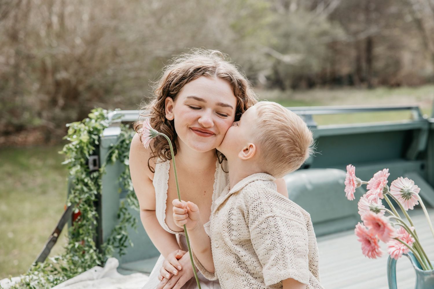 A young woman smiles as a toddler boy gives her a sweet kiss on the cheek outdoors near a decorated truck.