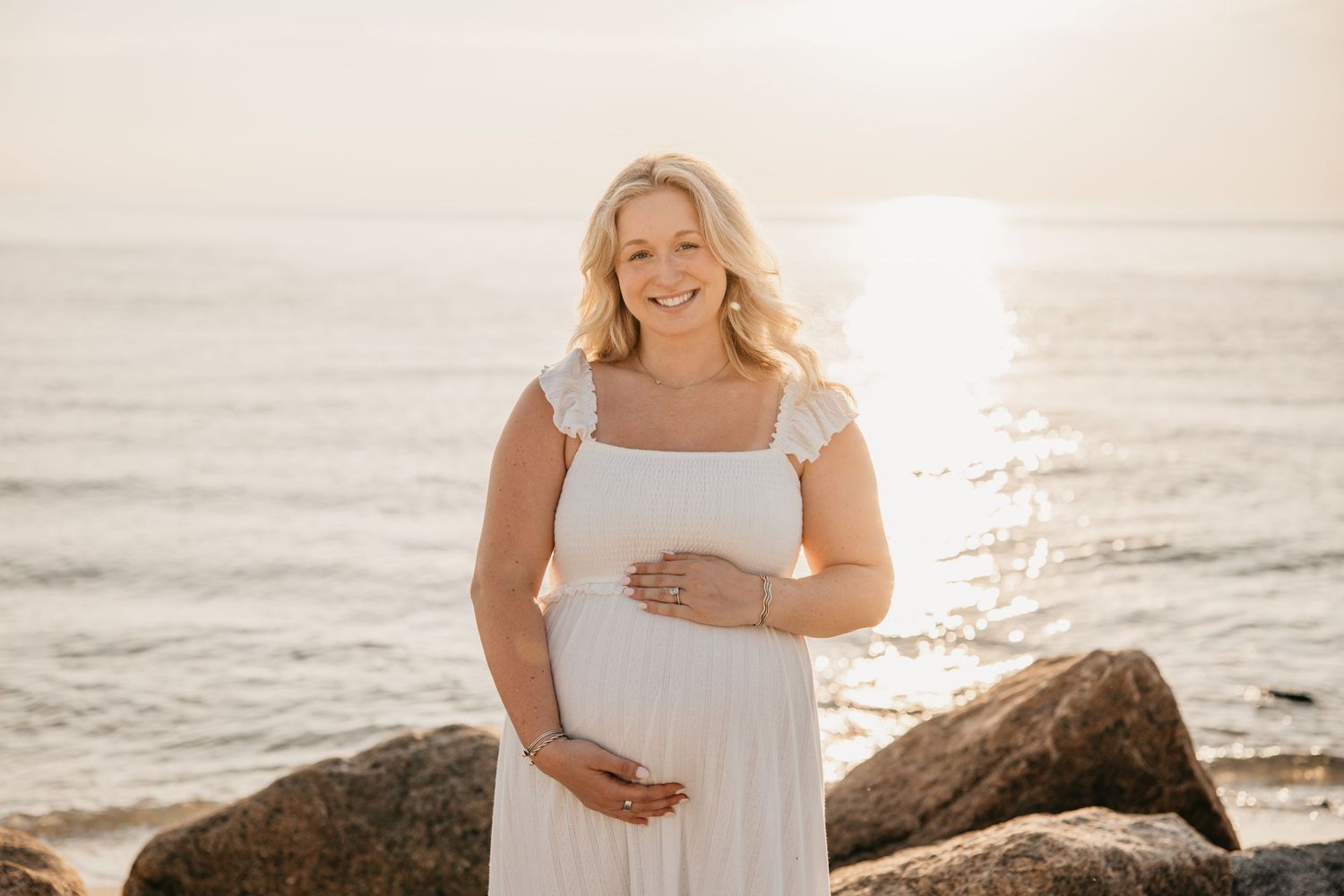 Pregnant woman in white dress cradling baby bump on rocky beach at golden sunset.