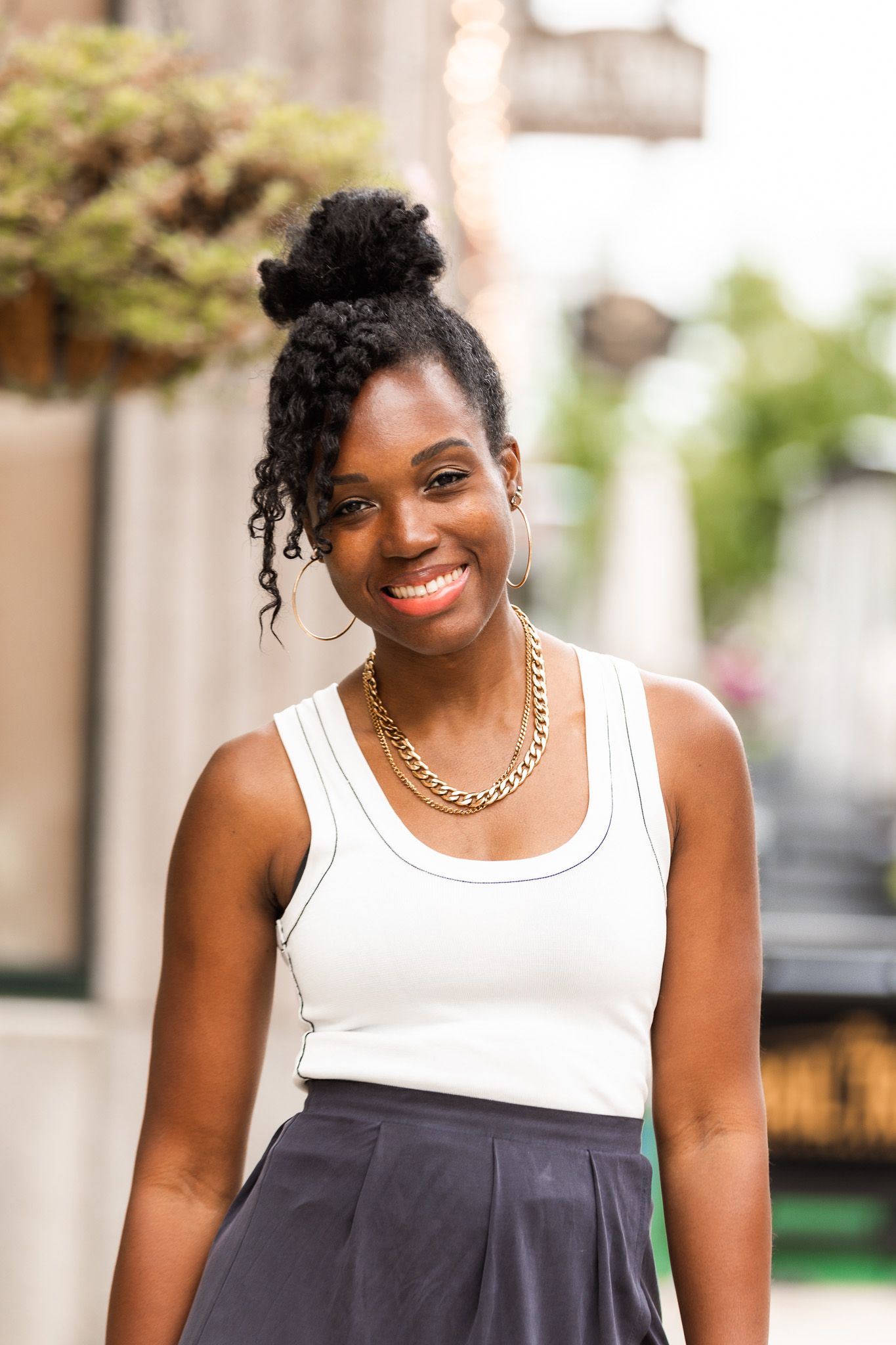 A person in a white tank top and dark skirt smiles warmly in an outdoor setting.