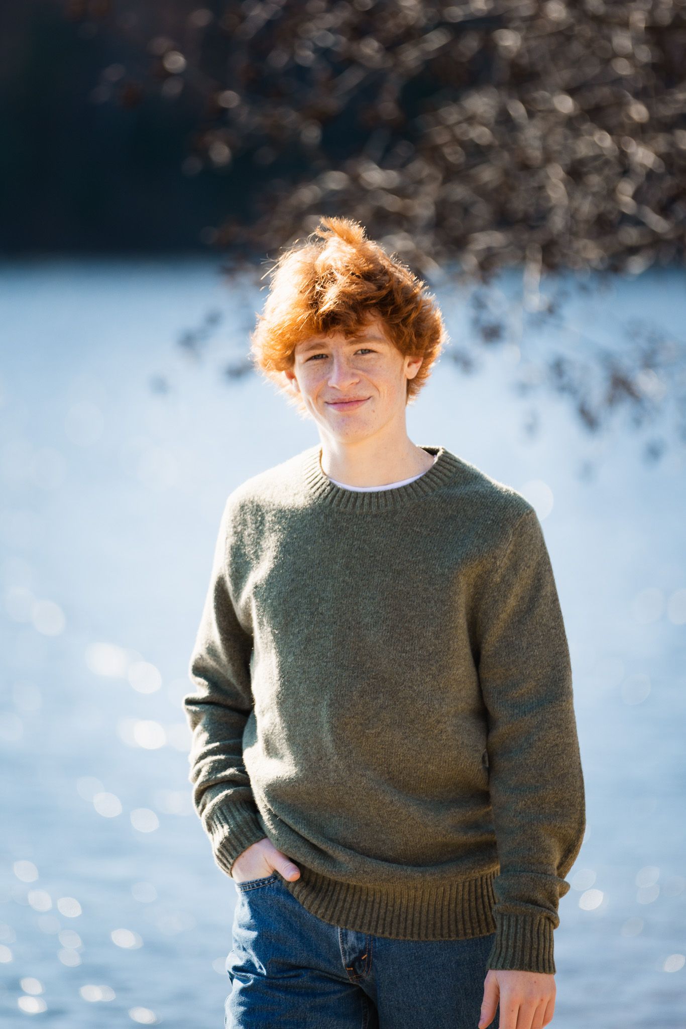 Person in casual winter attire poses near a snowy landscape with sunlit water in background.