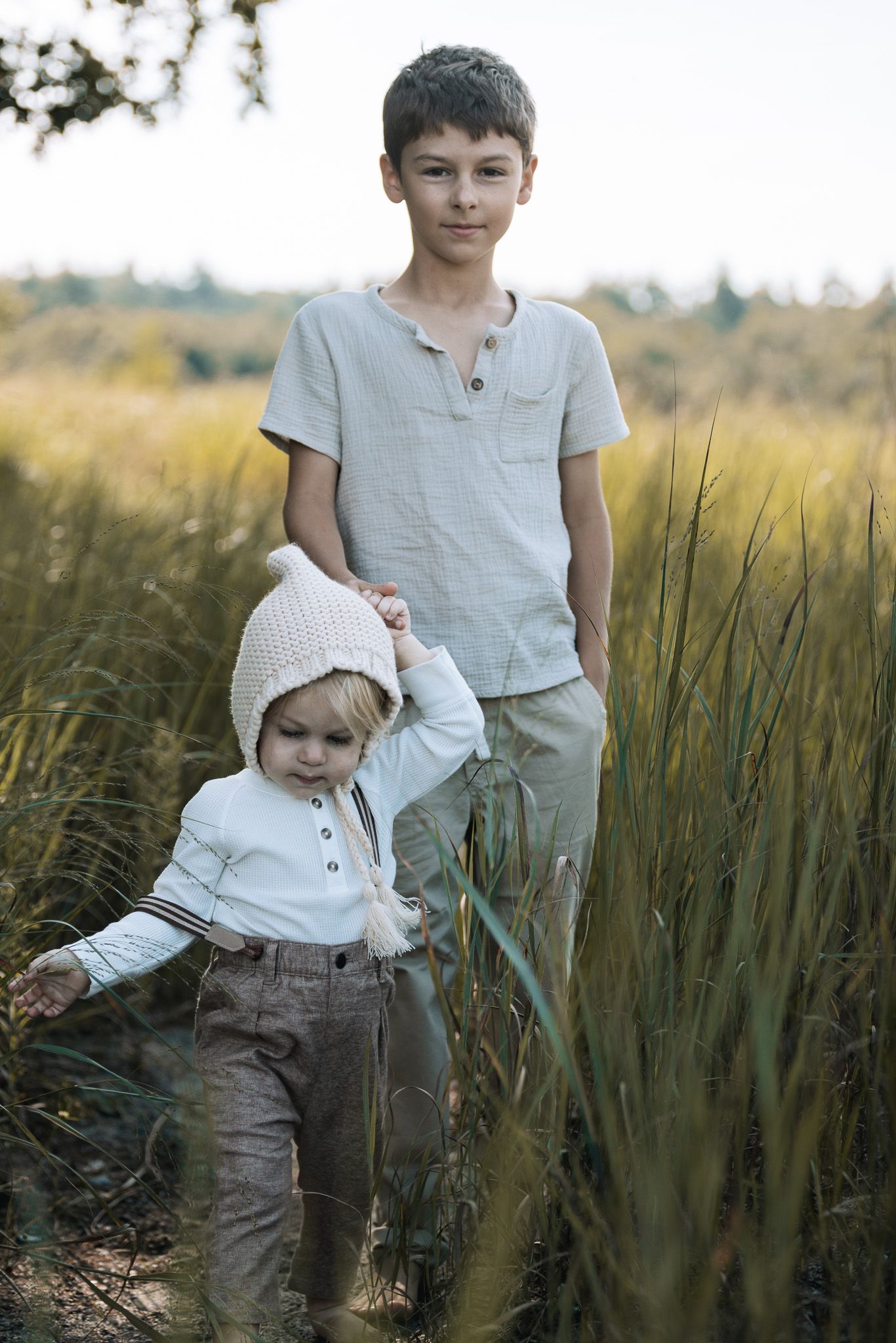 Two children explore a golden wheat field at sunset wearing light colored clothing.