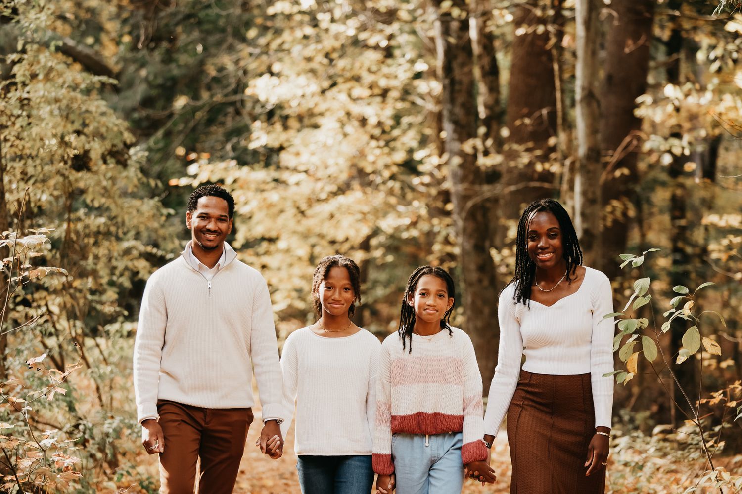 A family wearing coordinated white and brown outfits poses together in a wooded outdoor setting during autumn.