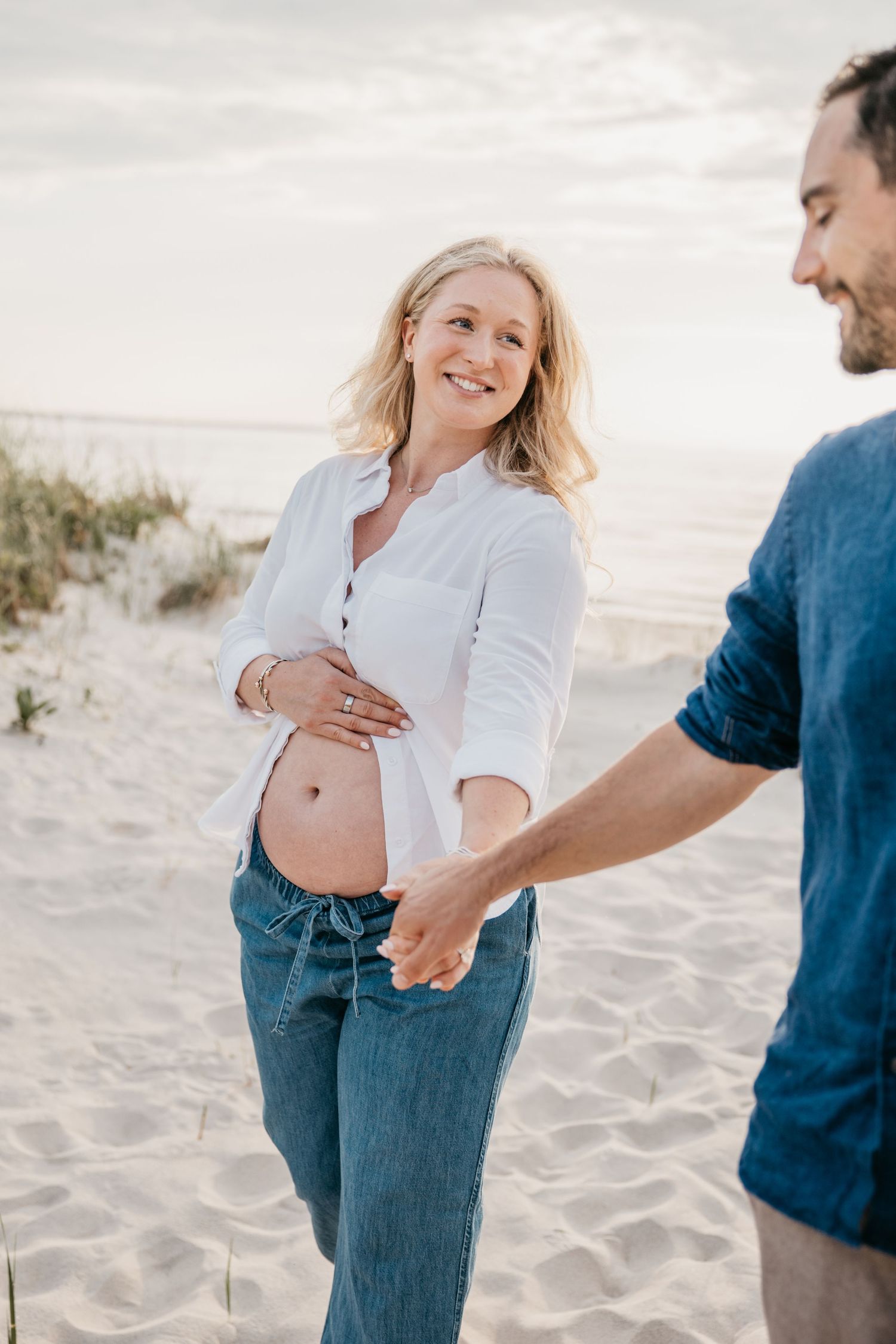 Pregnant woman in white shirt holds hands with partner on sandy beach, smiling joyfully during maternity photoshoot.