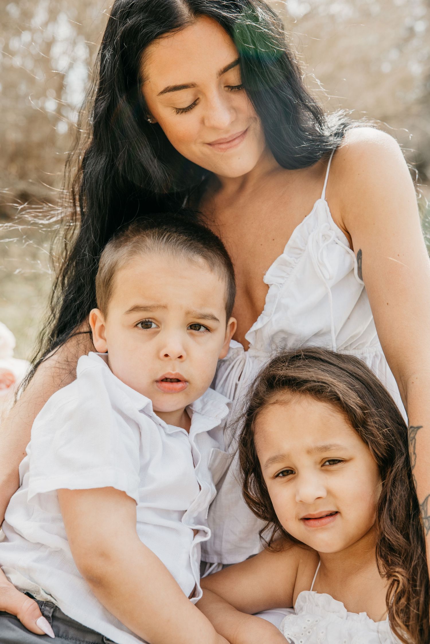 A woman in a white dress smiles warmly while embracing two young children outdoors in a sunny park.