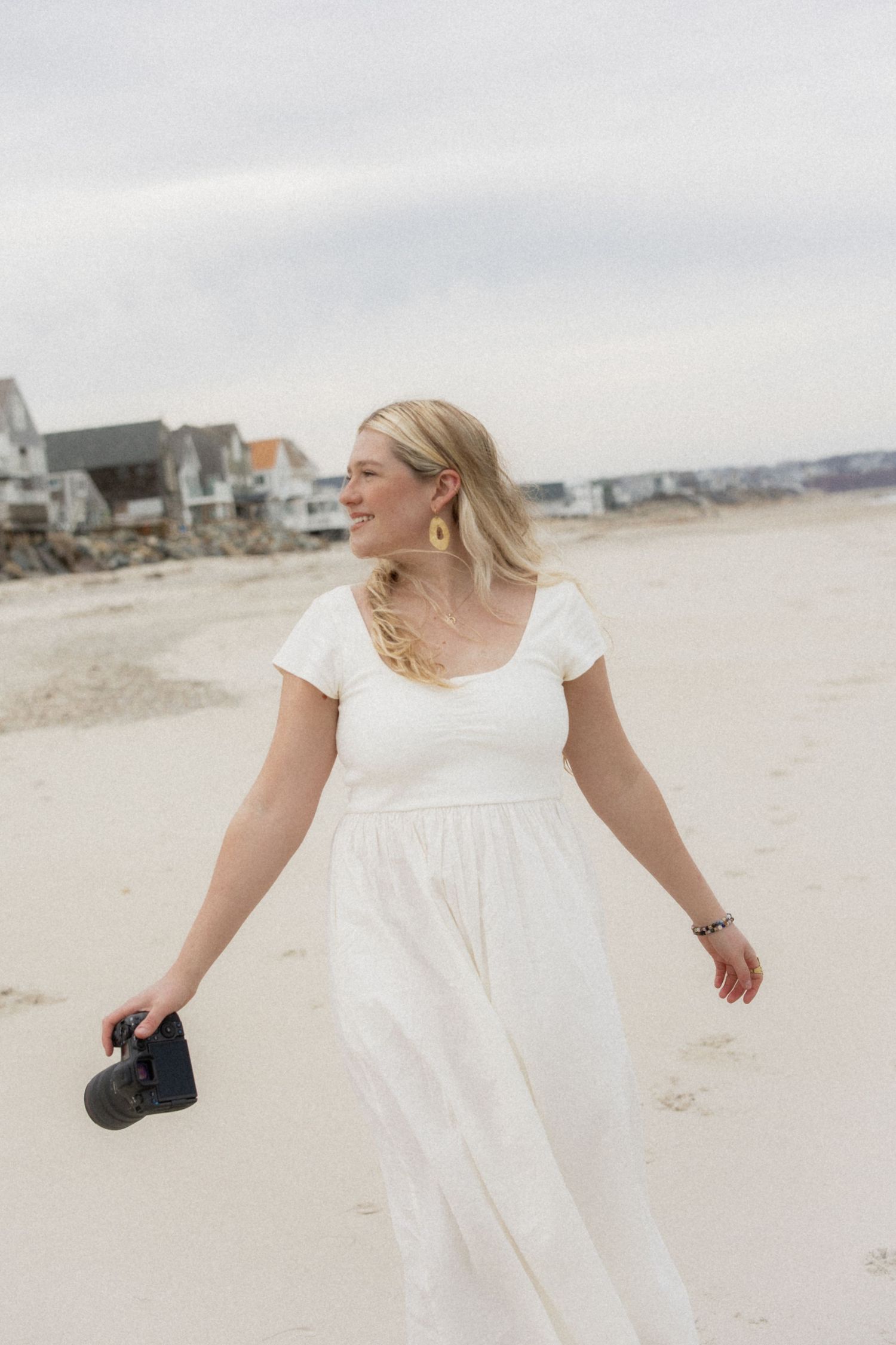 A person in a flowing white dress walks along a sandy beach on an overcast day.