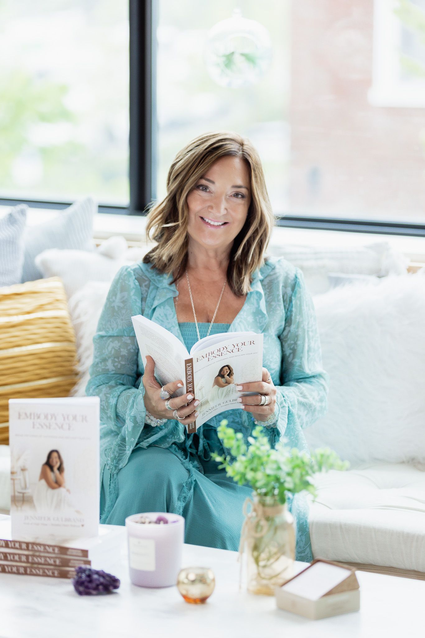 Woman in turquoise dress reading a book on cozy window seat with pillows.