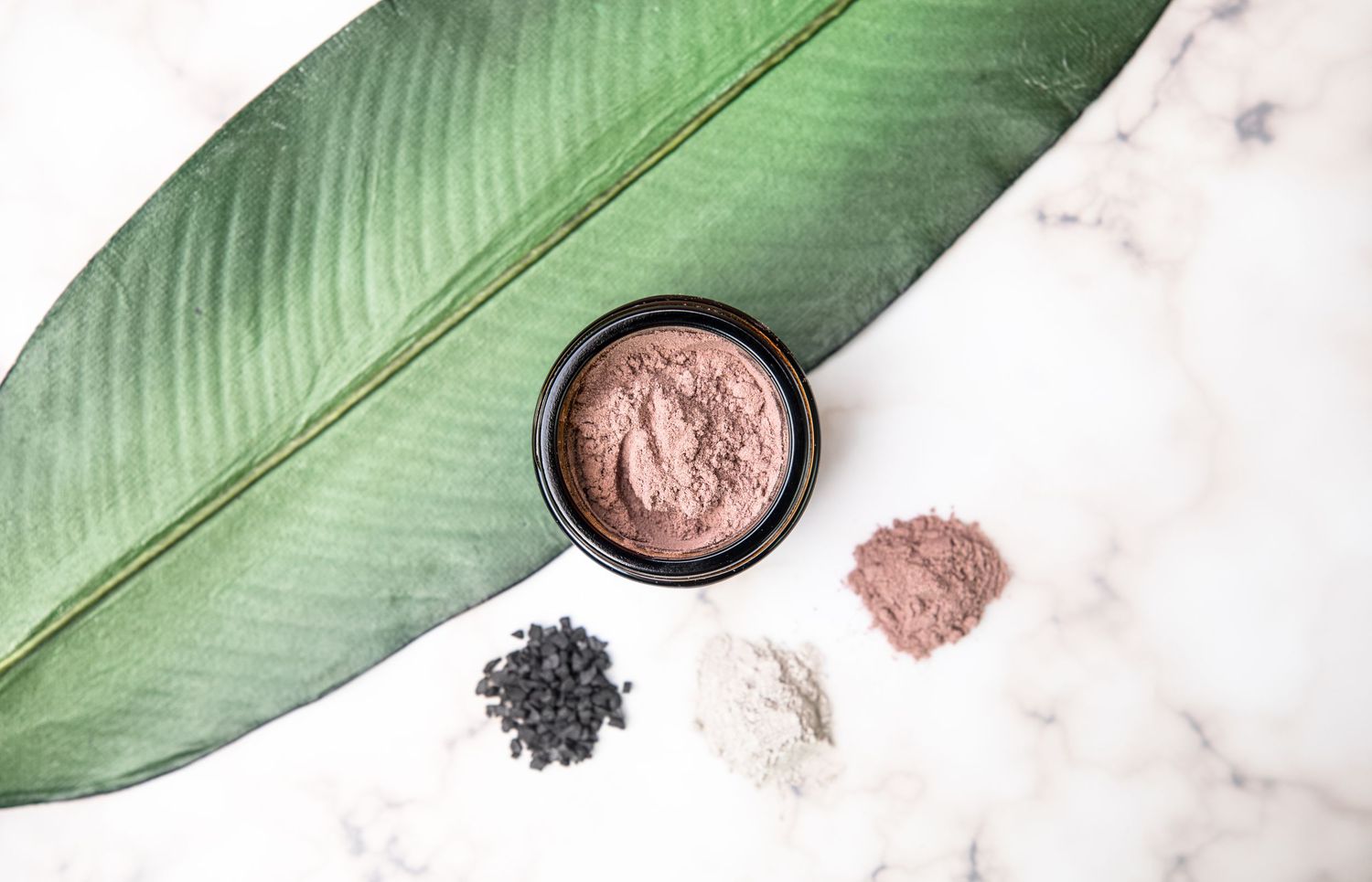 Pink clay powder and other natural ingredients displayed on marble surface with green leaf.