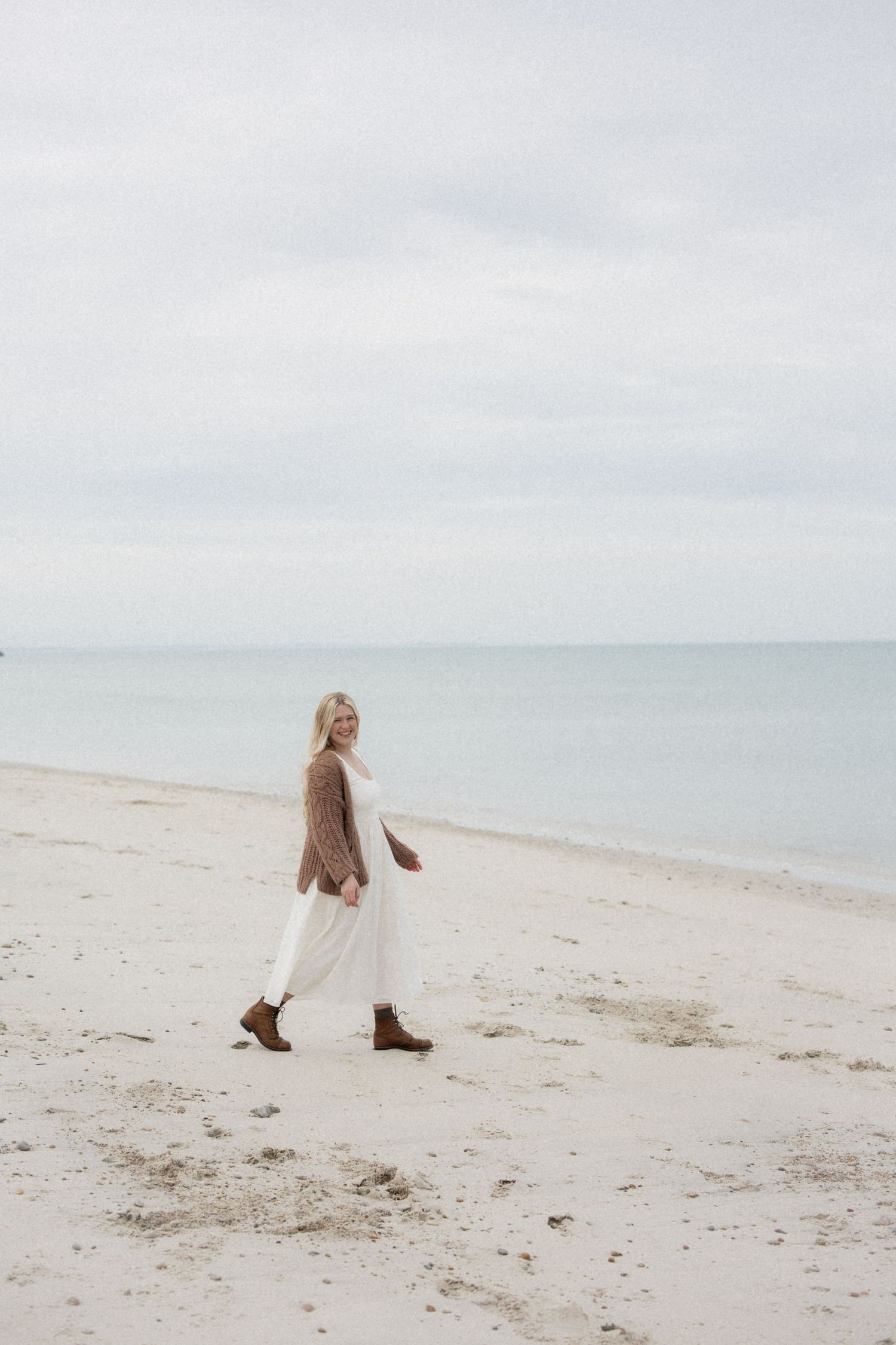 Person in brown sweater and white dress walks along serene beach shoreline on overcast day.
