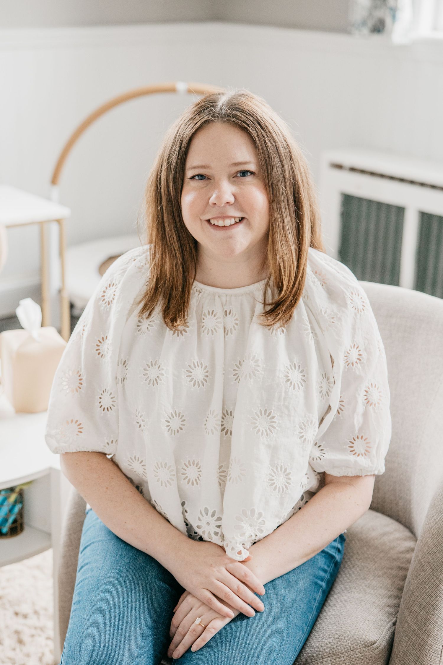 Smiling woman with brown hair wearing a white eyelet top and jeans, sitting in a cozy chair in a bright room.