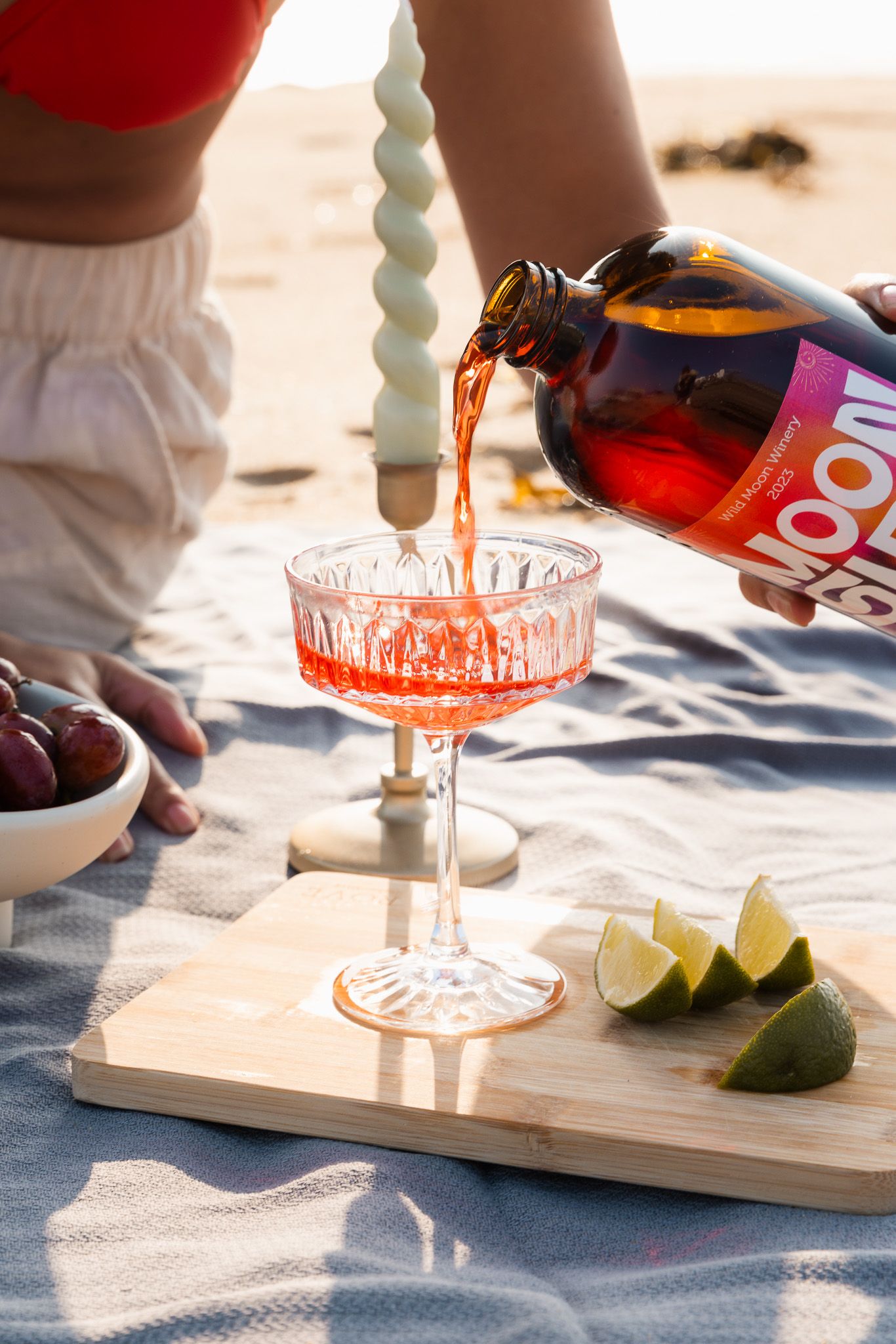 Pouring orange cocktail into glass with lime garnish on wooden serving board.