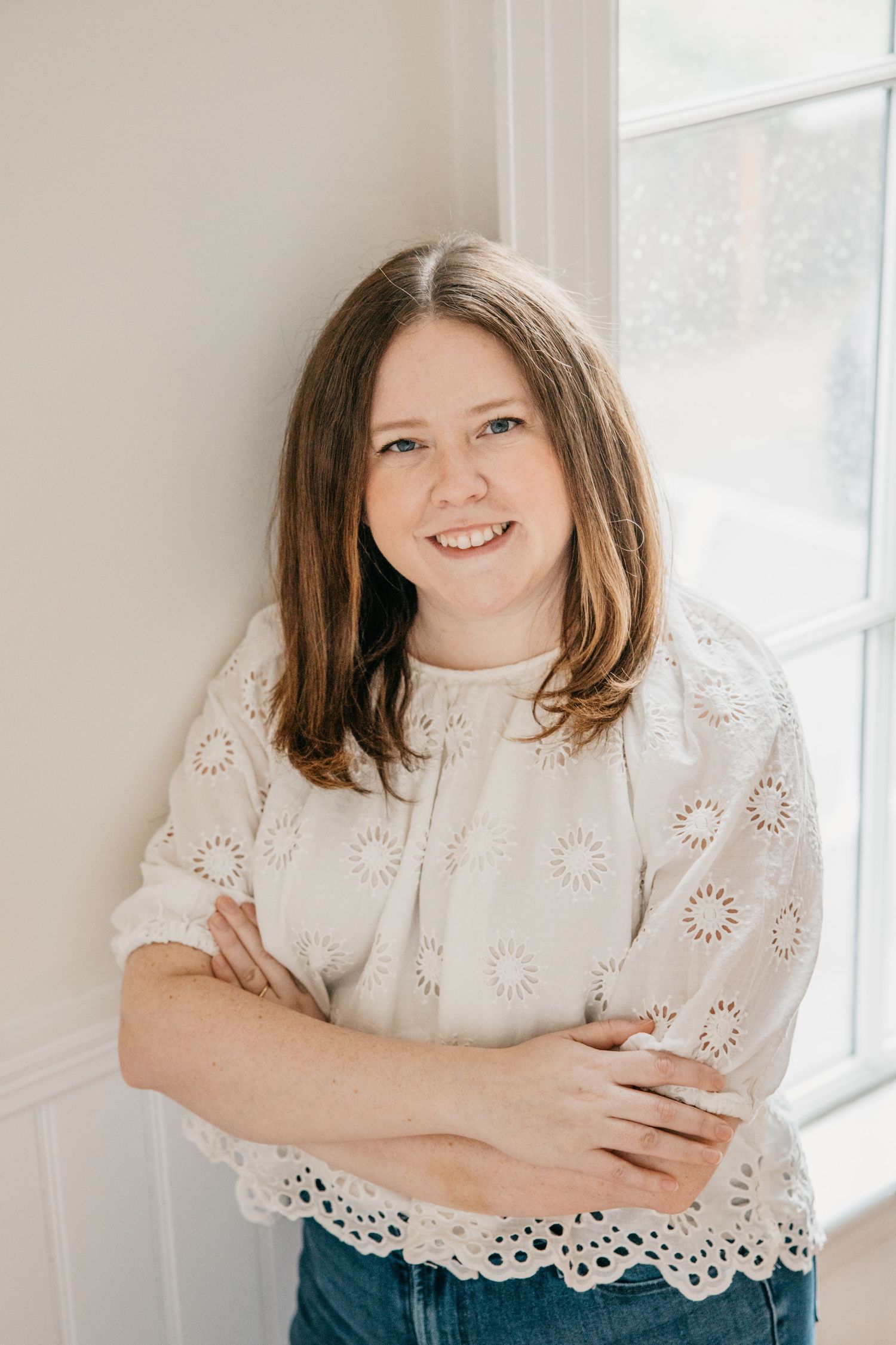 Smiling woman with brown hair in white eyelet top and jeans, arms crossed, leaning against wall near window.